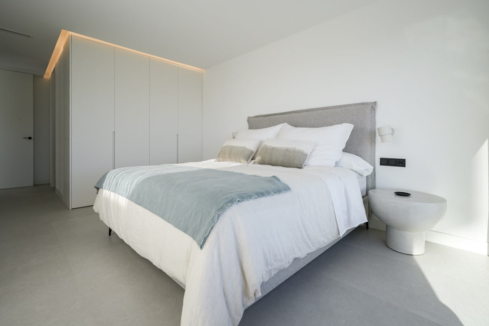 Dormitorio luminoso con cama doble, ropa de cama blanca y armario empotrado con iluminación superior.
