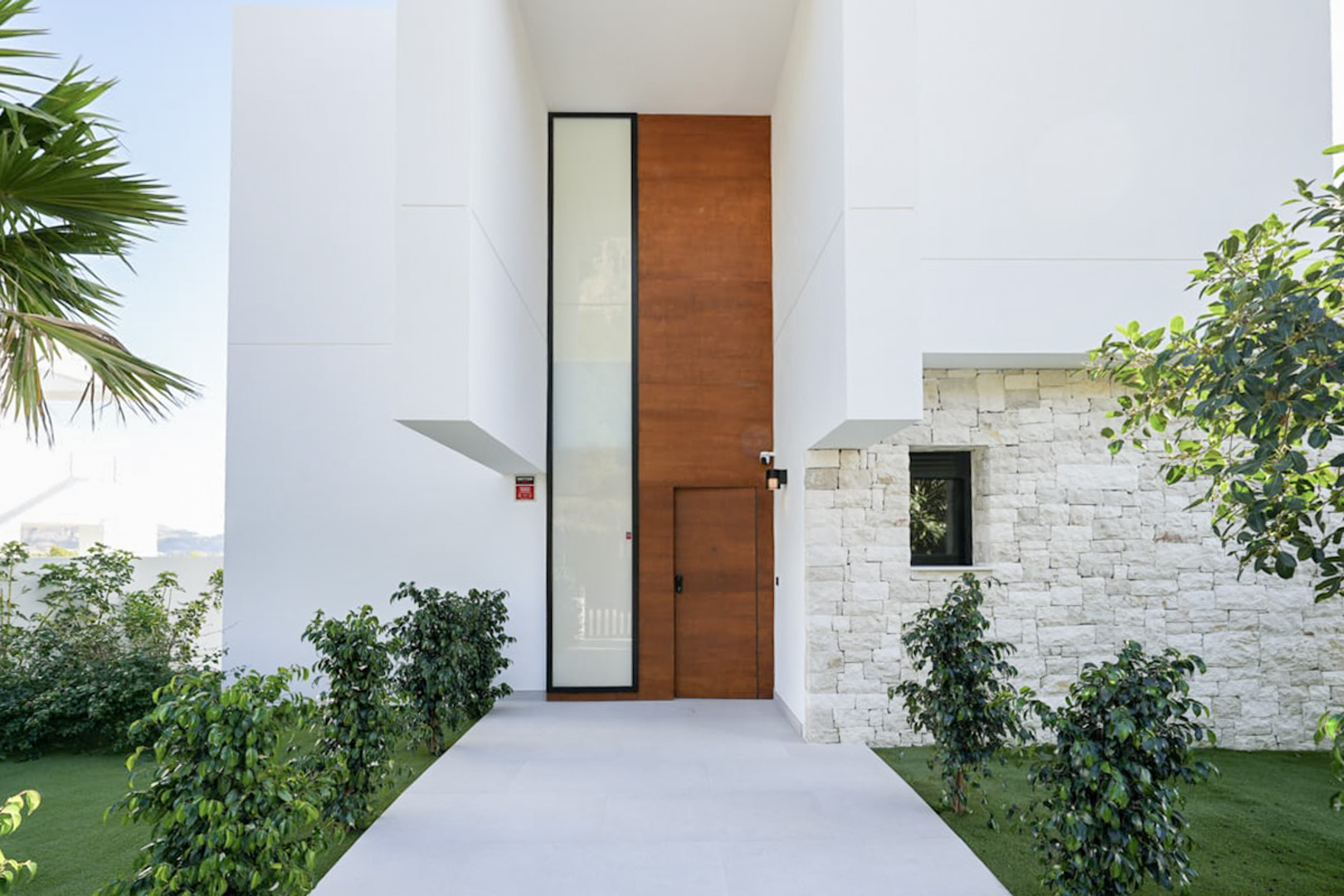 Fachada moderna con puerta de madera y detalles en piedra clara, rodeada de vegetación.