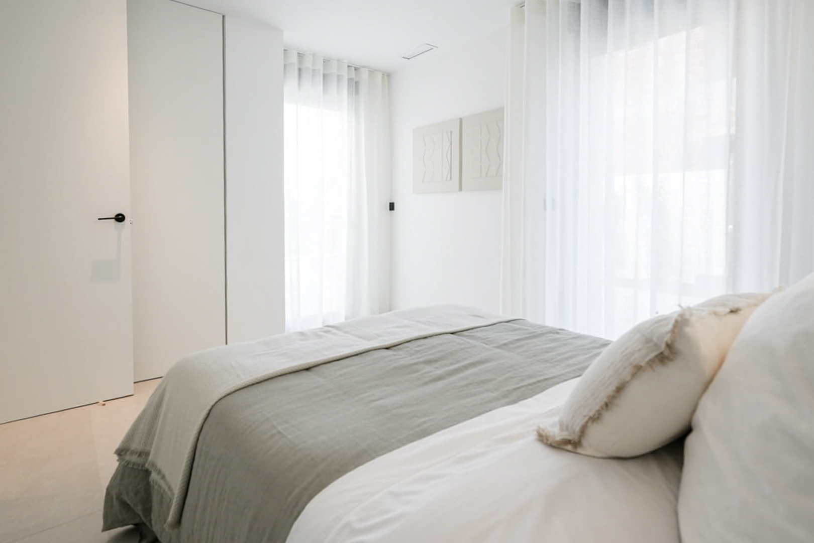Habitación luminosa con ropa de cama gris y blanca, cortinas traslúcidas y decoración minimalista.
