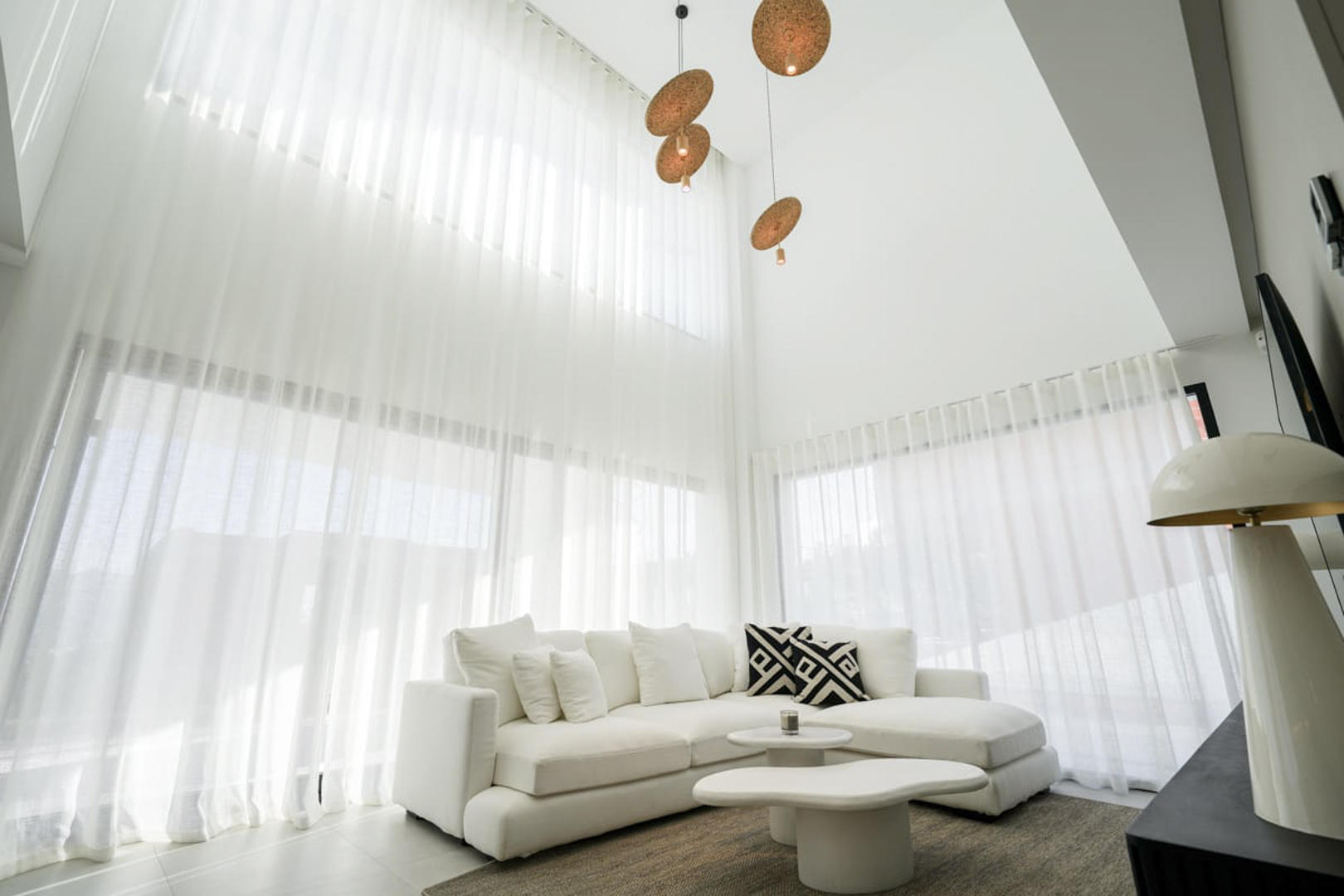 Salón luminoso con cortinas blancas, sofá esquinero y lámparas colgantes de diseño, creando un ambiente moderno y minimalista.