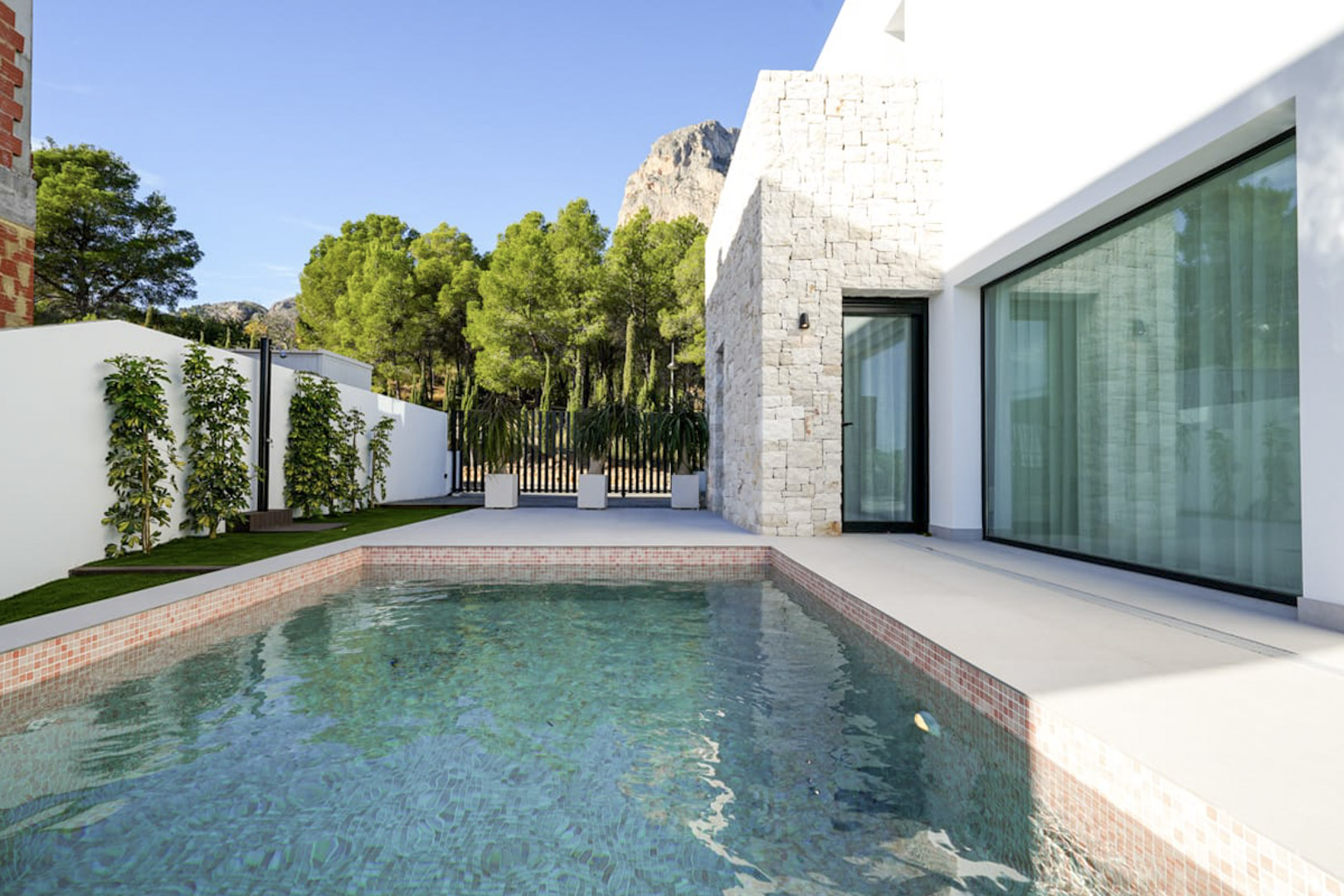 Vista de la piscina privada con borde de mosaico y fachada de piedra blanca, rodeada de vegetación exuberante.