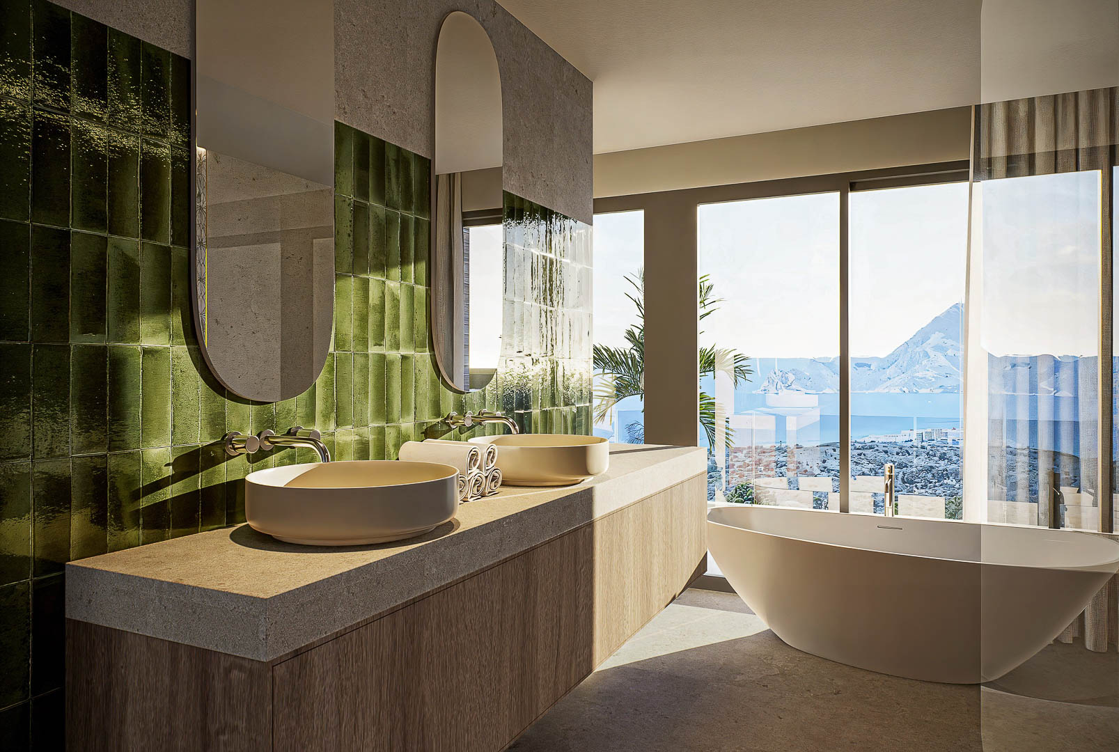 Baño principal con lavabos dobles sobre encimera de piedra, azulejos verdes verticales y bañera exenta frente a ventanales con vistas al mar y al Peñón de Ifach.