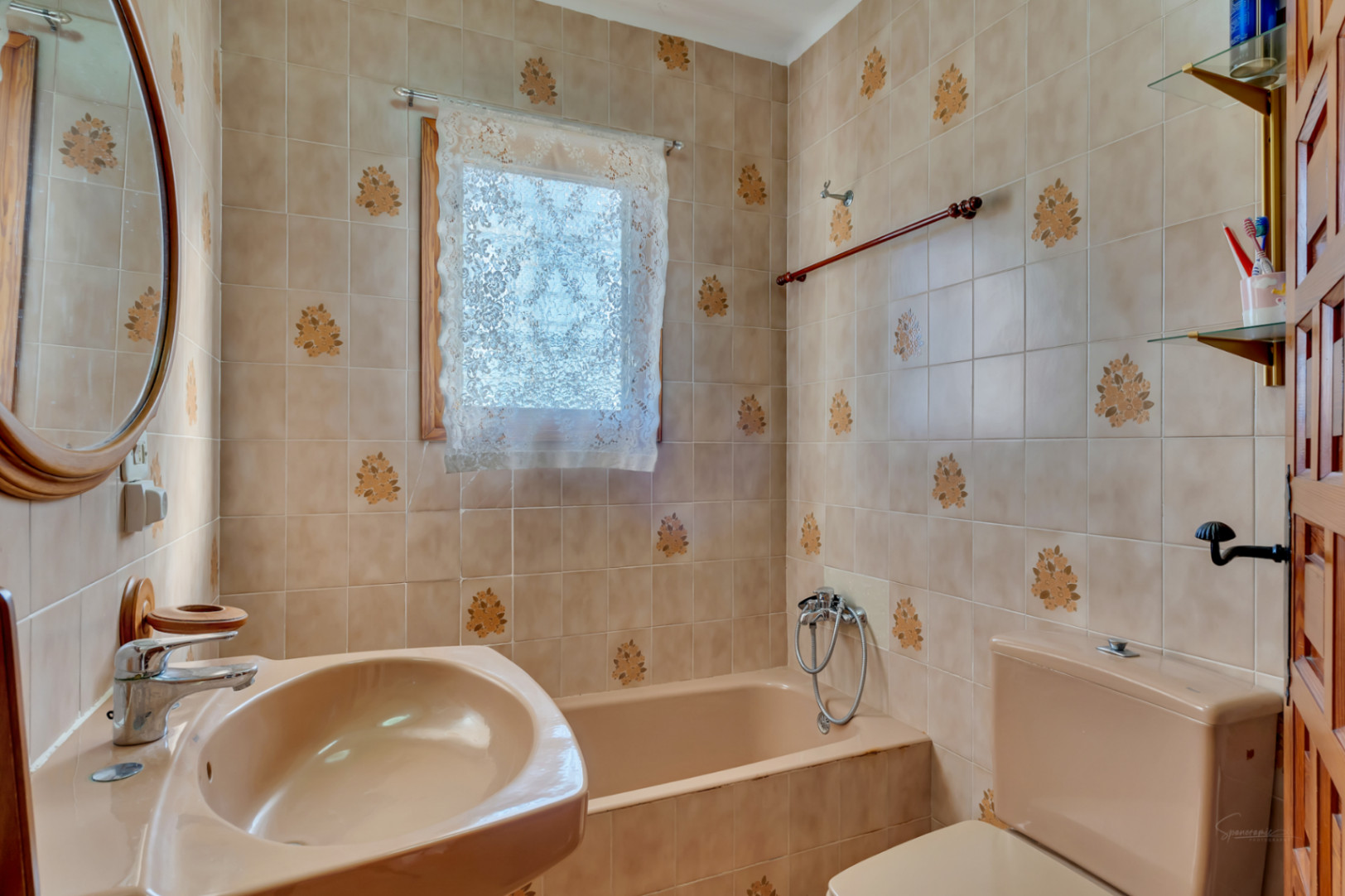 Baño de estilo tradicional con azulejos beige decorados, bañera integrada y lavabo a juego. Incluye detalles en madera, espejo ovalado y una ventana con cortina de encaje.