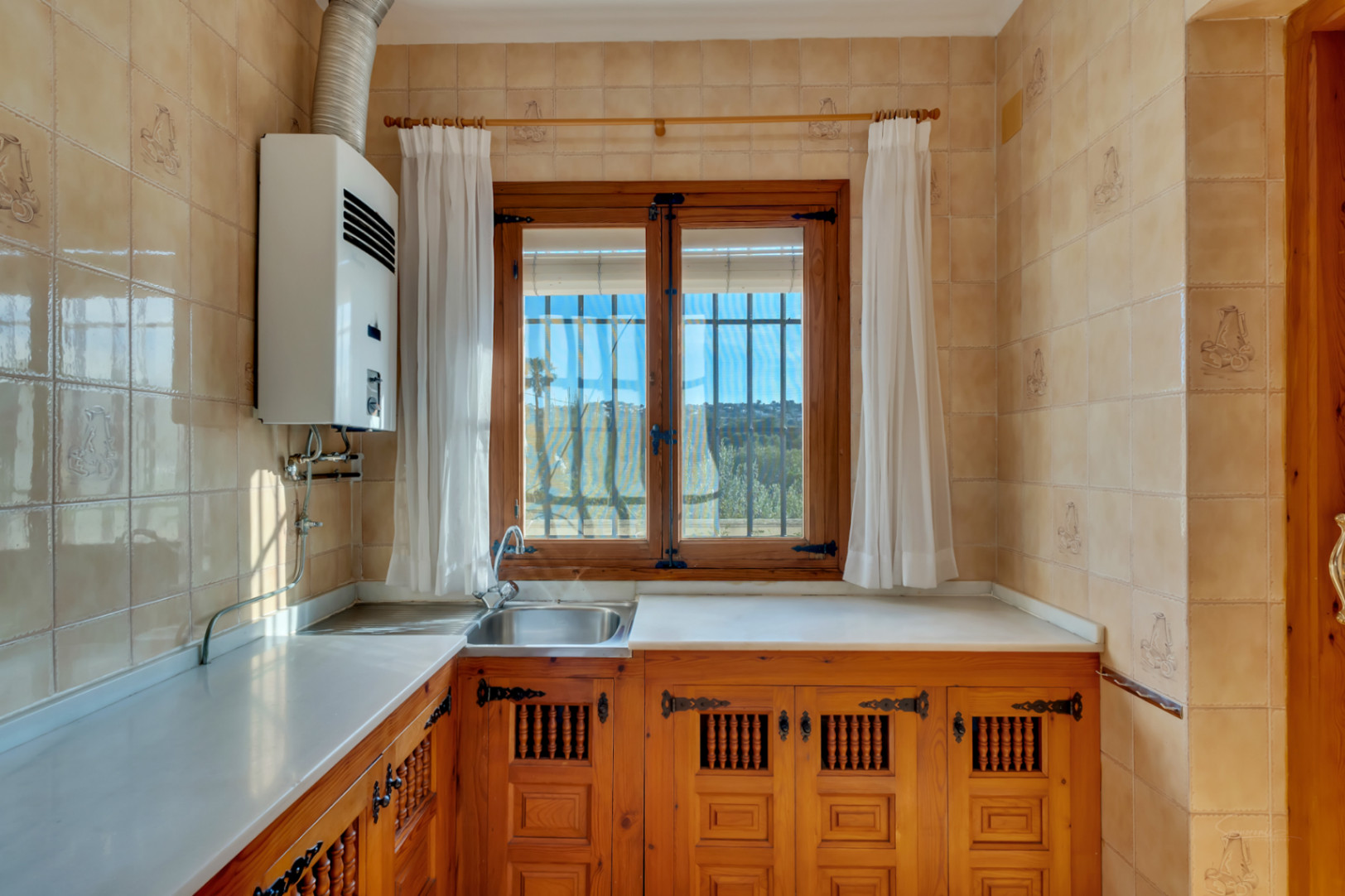 Cocina de estilo rústico con armarios de madera tallada, encimera blanca y ventana de madera. Incluye fregadero de acero inoxidable y calentador de gas de pared.