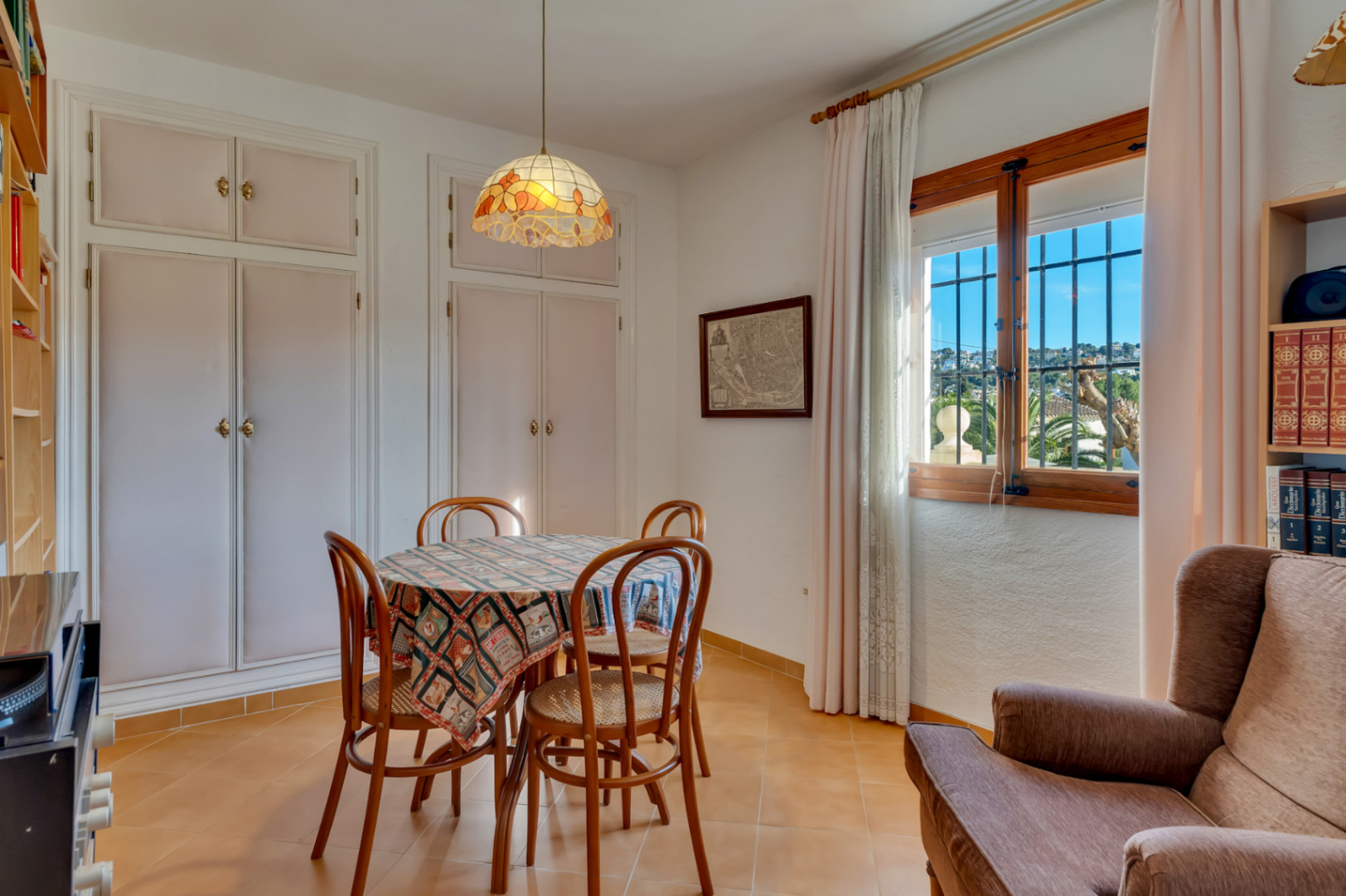 Comedor luminoso con mesa redonda, sillas de madera y lámpara de estilo Tiffany. Incluye armarios empotrados blancos y ventana con vistas al entorno de Jávea.