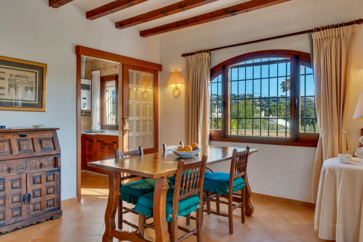 Comedor tradicional con vigas de madera a la vista, mesa de madera maciza y acceso a la cocina mediante puerta corredera. Destaca el ventanal en arco con vistas al exterior.