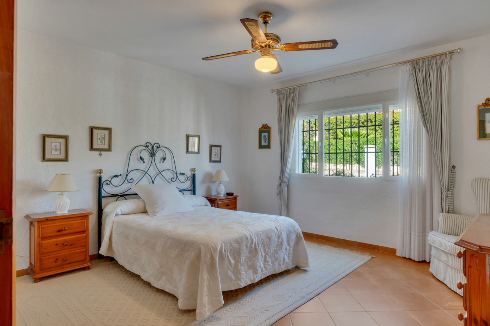 Dormitorio luminoso con cabecero de forja, suelos de gres cerámico y ventilador de techo. La ventana ofrece vistas al jardín y luz natural constante.