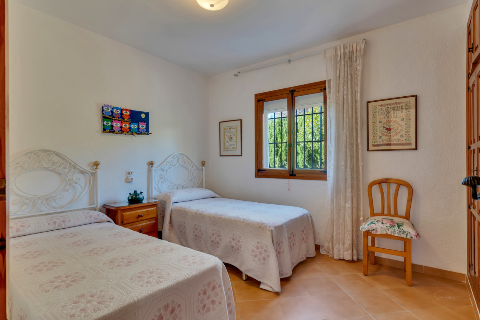 Dormitorio luminoso con dos camas individuales, cabeceros de forja blanca, suelos cerámicos y ventana de madera con vistas al exterior.