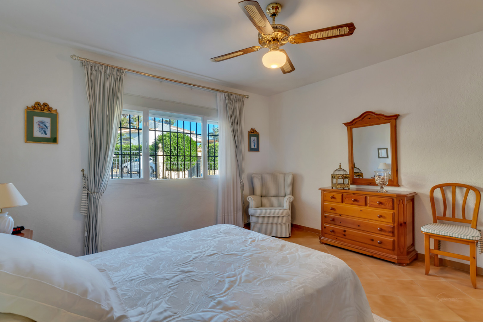 Dormitorio luminoso con suelos de terracota, mobiliario de madera tradicional, ventilador de techo y ventana con vistas al jardín exterior.