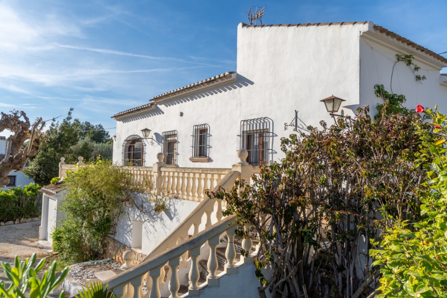 Fachada blanca de estilo mediterráneo con balaustrada clásica, ventanas protegidas por rejas de hierro forjado y una escalera exterior que conduce a la terraza principal.