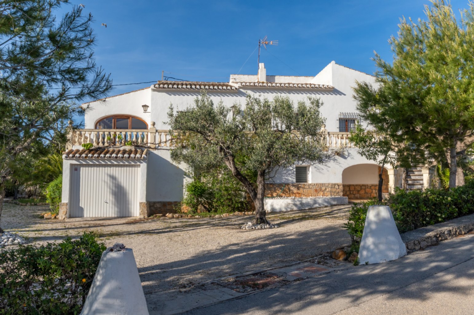Fachada de villa mediterránea con muros blancos, terraza con balaustrada y garaje. El exterior cuenta con un olivo central y base de piedra natural en una parcela llana.