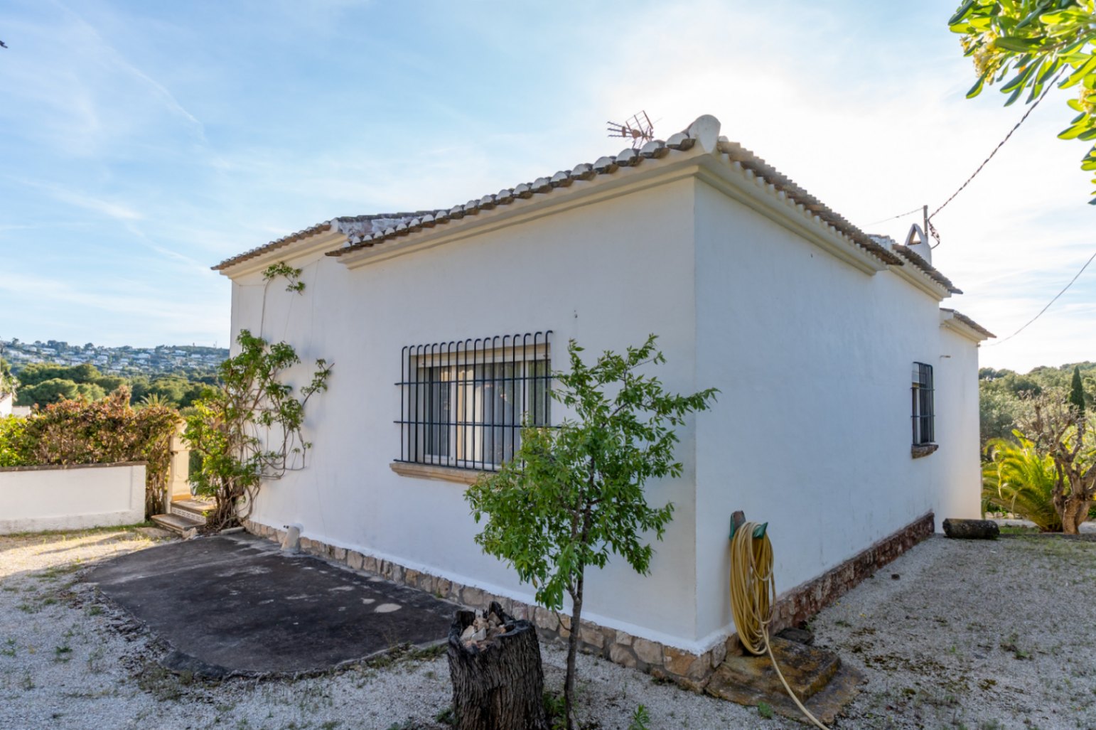 Fachada exterior de la villa con paredes blancas, ventanas protegidas por rejas de hierro y base de piedra natural, rodeada de un terreno de grava y vegetación.