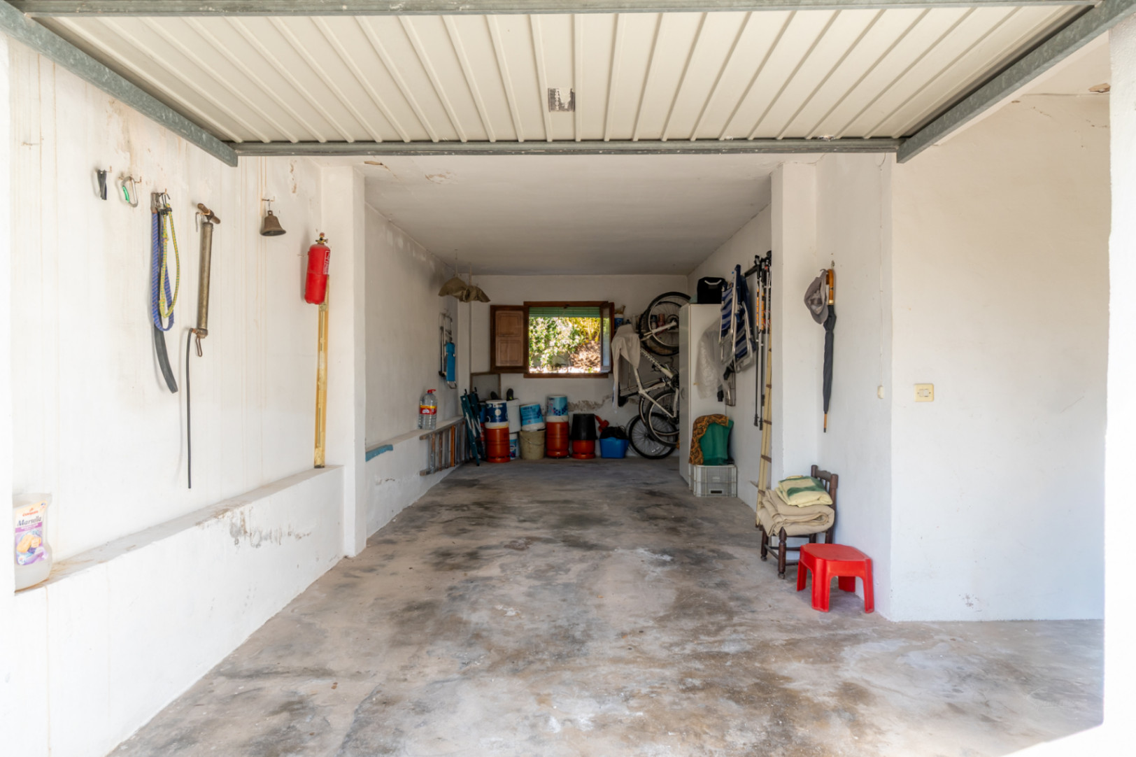 Garaje funcional con suelo de hormigón y paredes blancas, ofreciendo amplio espacio para almacenamiento y herramientas, con luz natural desde una ventana posterior.