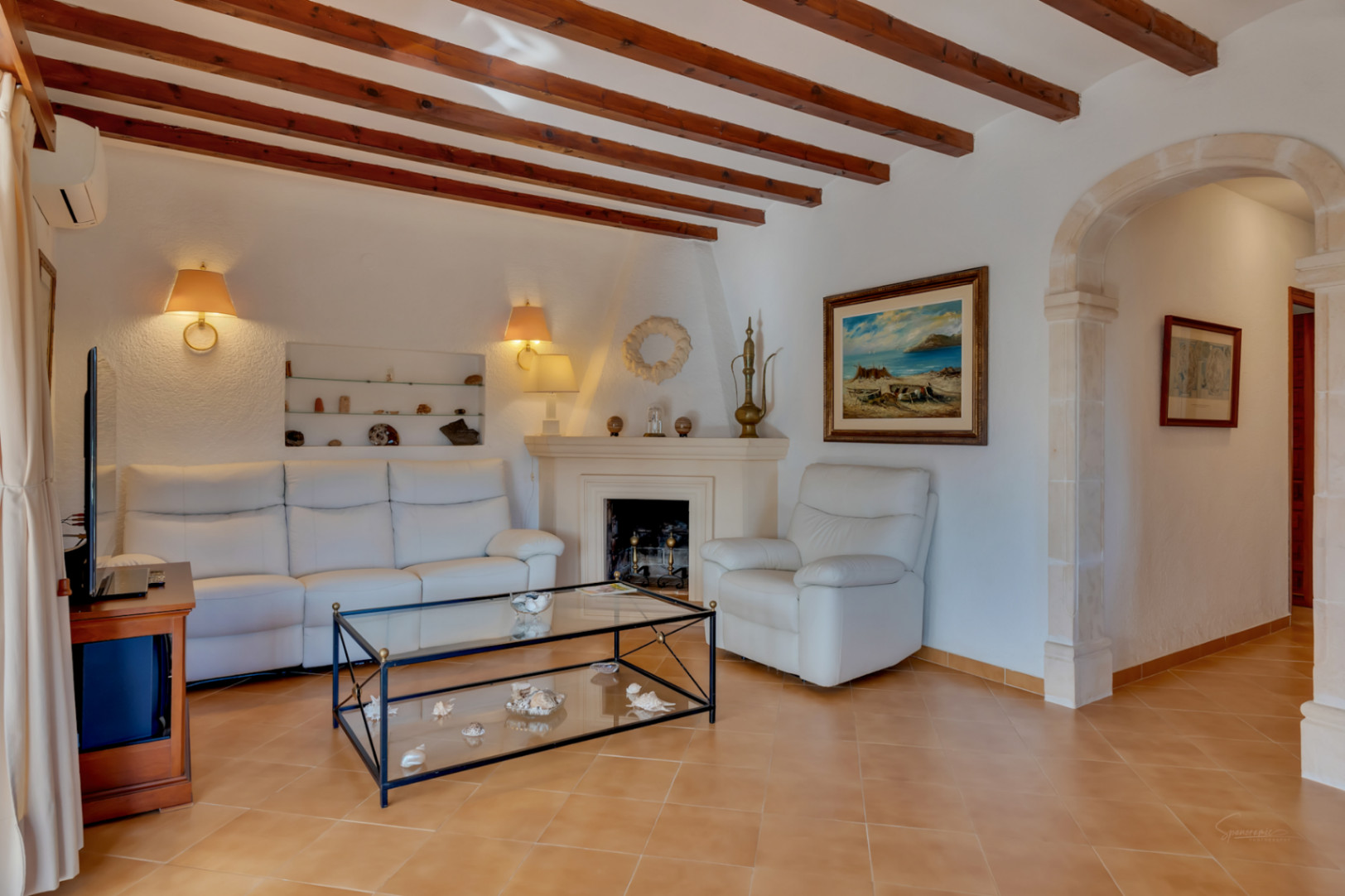 Salón con vigas de madera, chimenea de obra y un arco de piedra tosca tradicional. Incluye sofás de cuero blanco y suelos cerámicos en tonos cálidos.