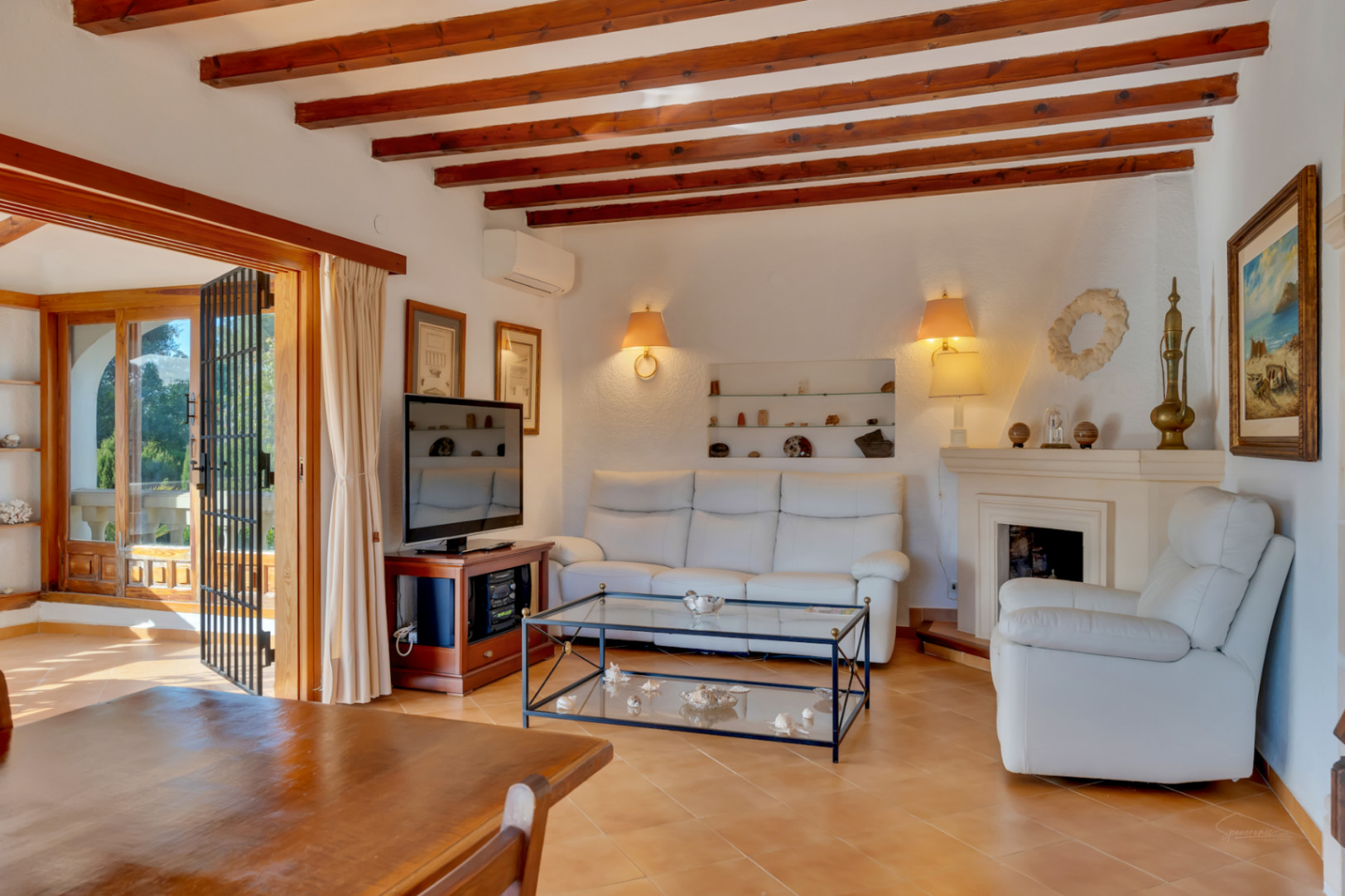 Salón con vigas de madera vista, chimenea clásica y sofás de cuero blanco. El espacio cuenta con suelos cerámicos y acceso directo a la terraza exterior.