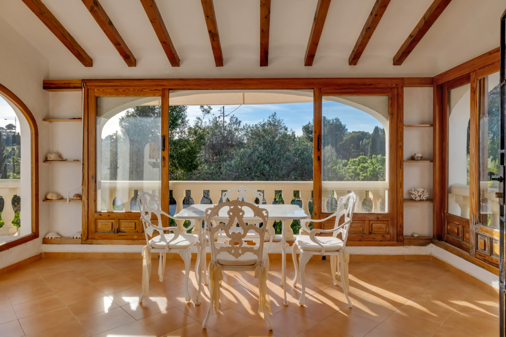 Terraza acristalada con vigas de madera, suelos de terracota y mobiliario de hierro forjado blanco, ofreciendo vistas al jardín mediterráneo.