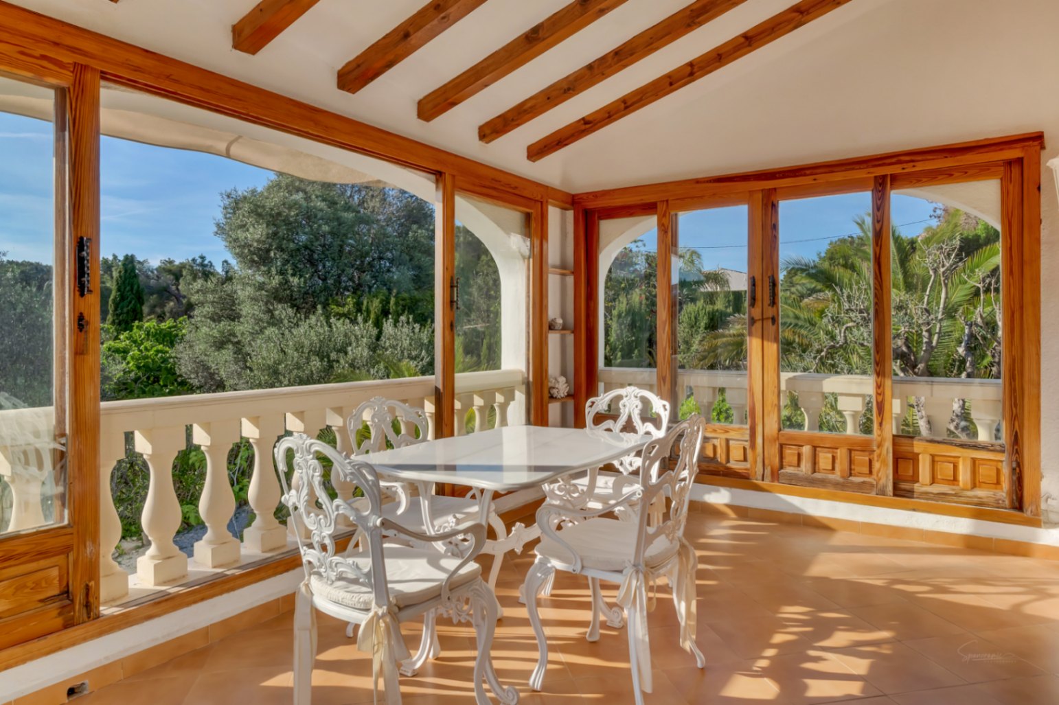 Terraza acristalada con vigas de madera vista, mobiliario de hierro forjado blanco y amplios ventanales con vistas al jardín mediterráneo.