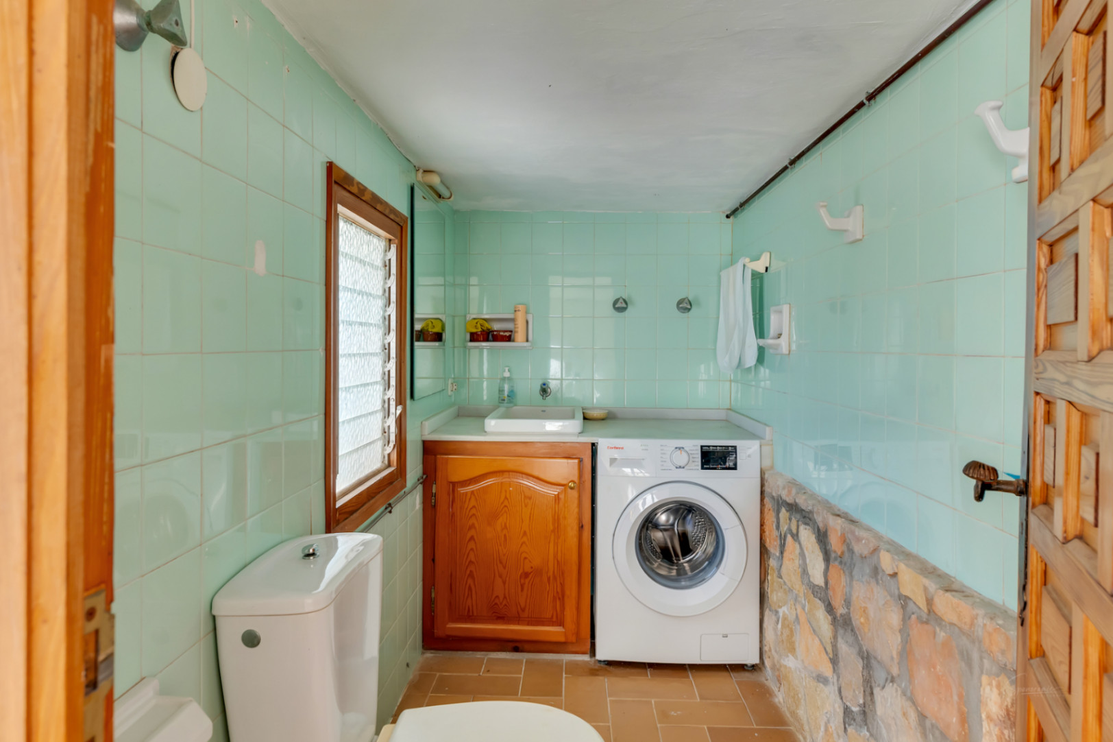 Zona de lavandería y aseo con paredes de azulejo verde, base de piedra natural, lavadora integrada y mueble de madera bajo el lavabo.