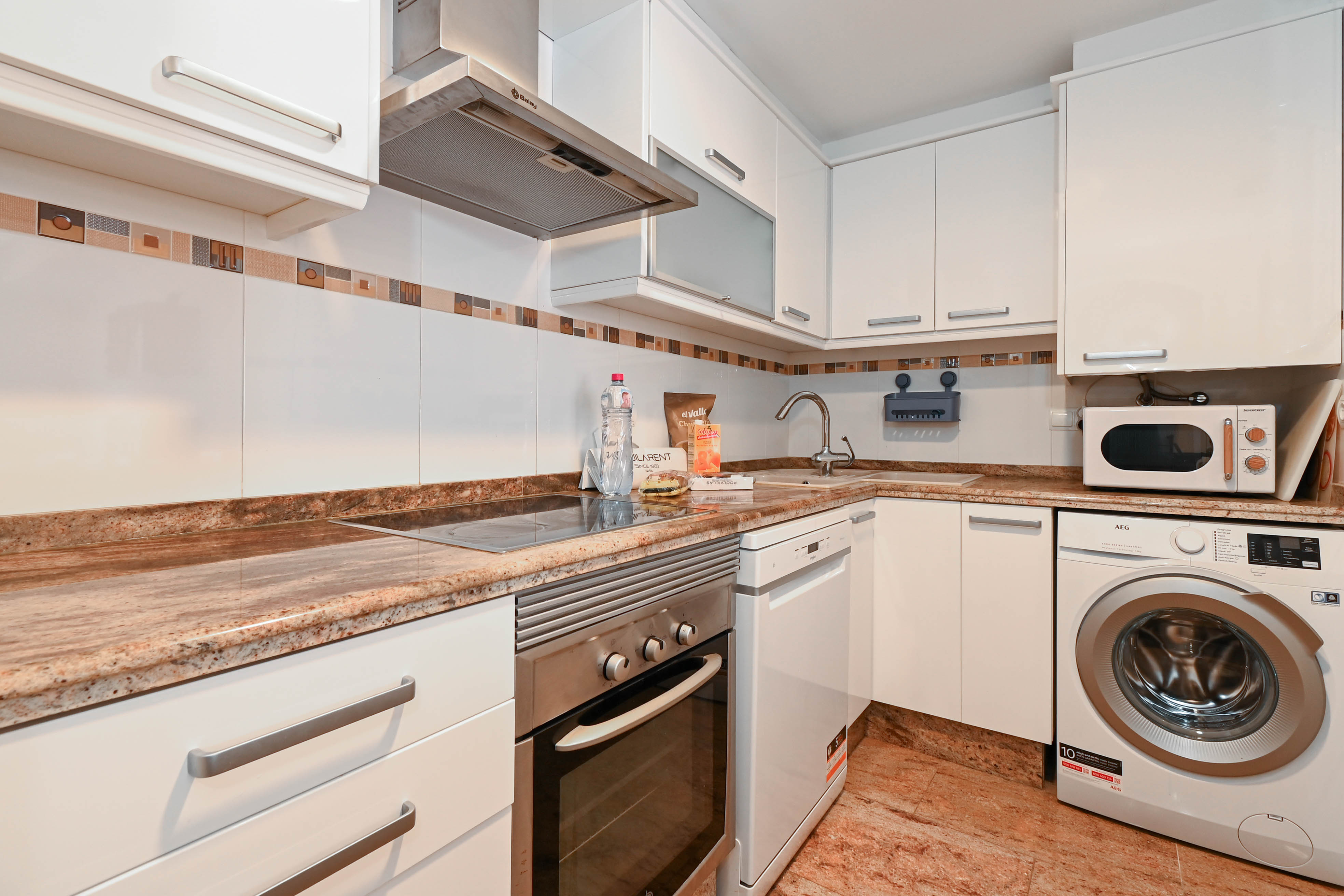 Cocina funcional equipada con armarios blancos, encimeras de granito veteado y electrodomésticos integrados como lavadora, lavavajillas y horno de acero inoxidable.
