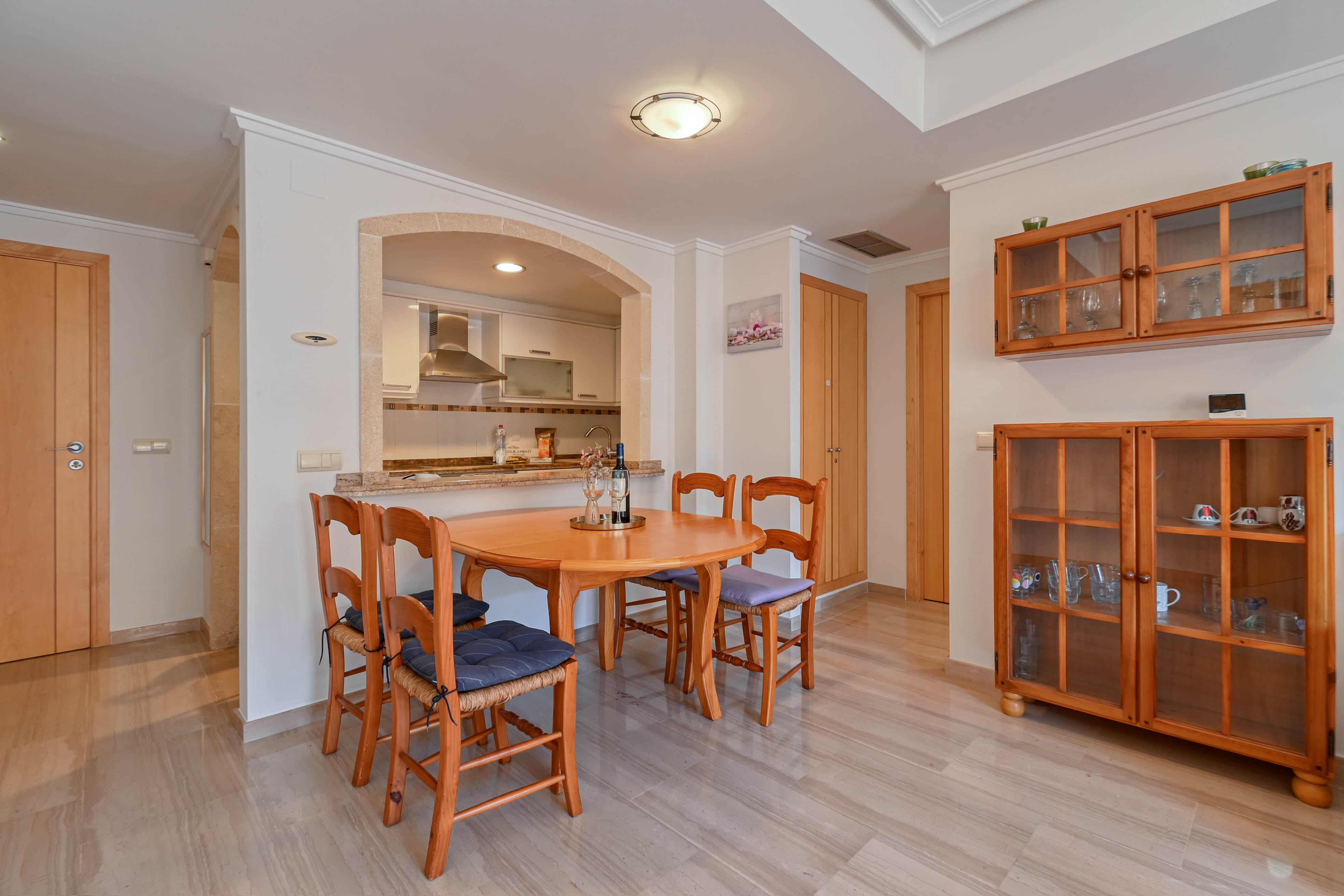 Comedor con mesa de madera y sillas, conectado a la cocina mediante un arco de piedra natural y suelos de mármol pulido.