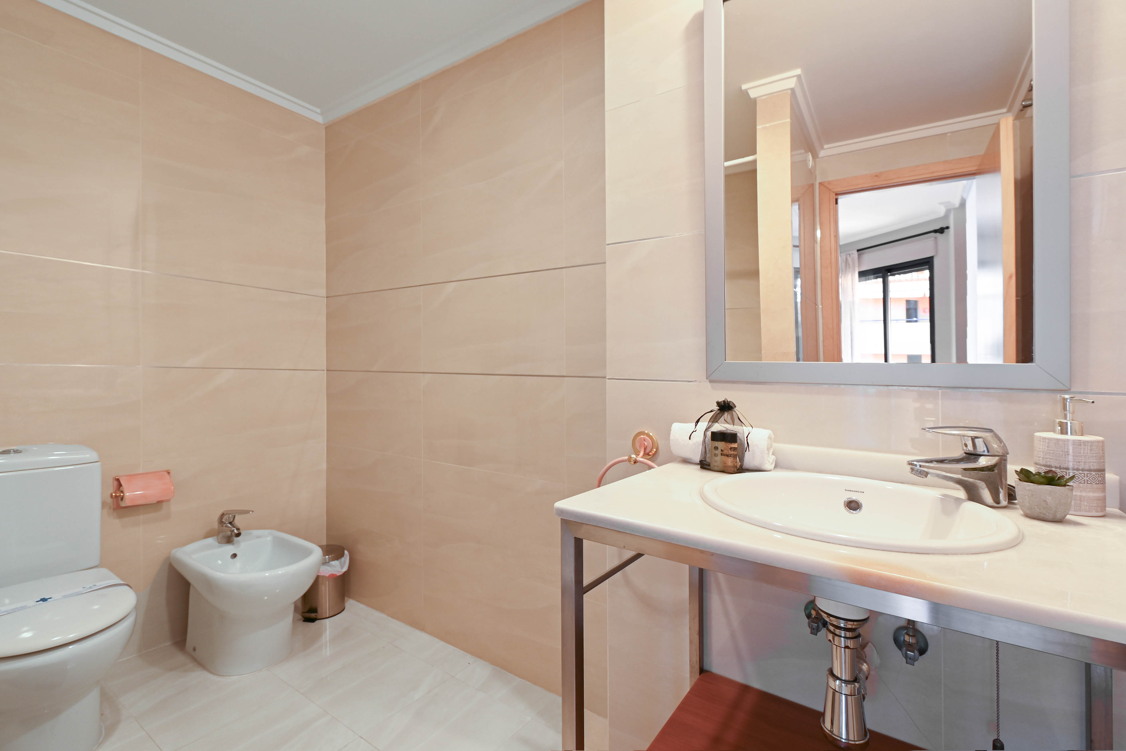 Cuarto de baño con paredes alicatadas en tonos beige, equipado con inodoro, bidé y lavabo encastrado sobre encimera de piedra con estructura metálica.