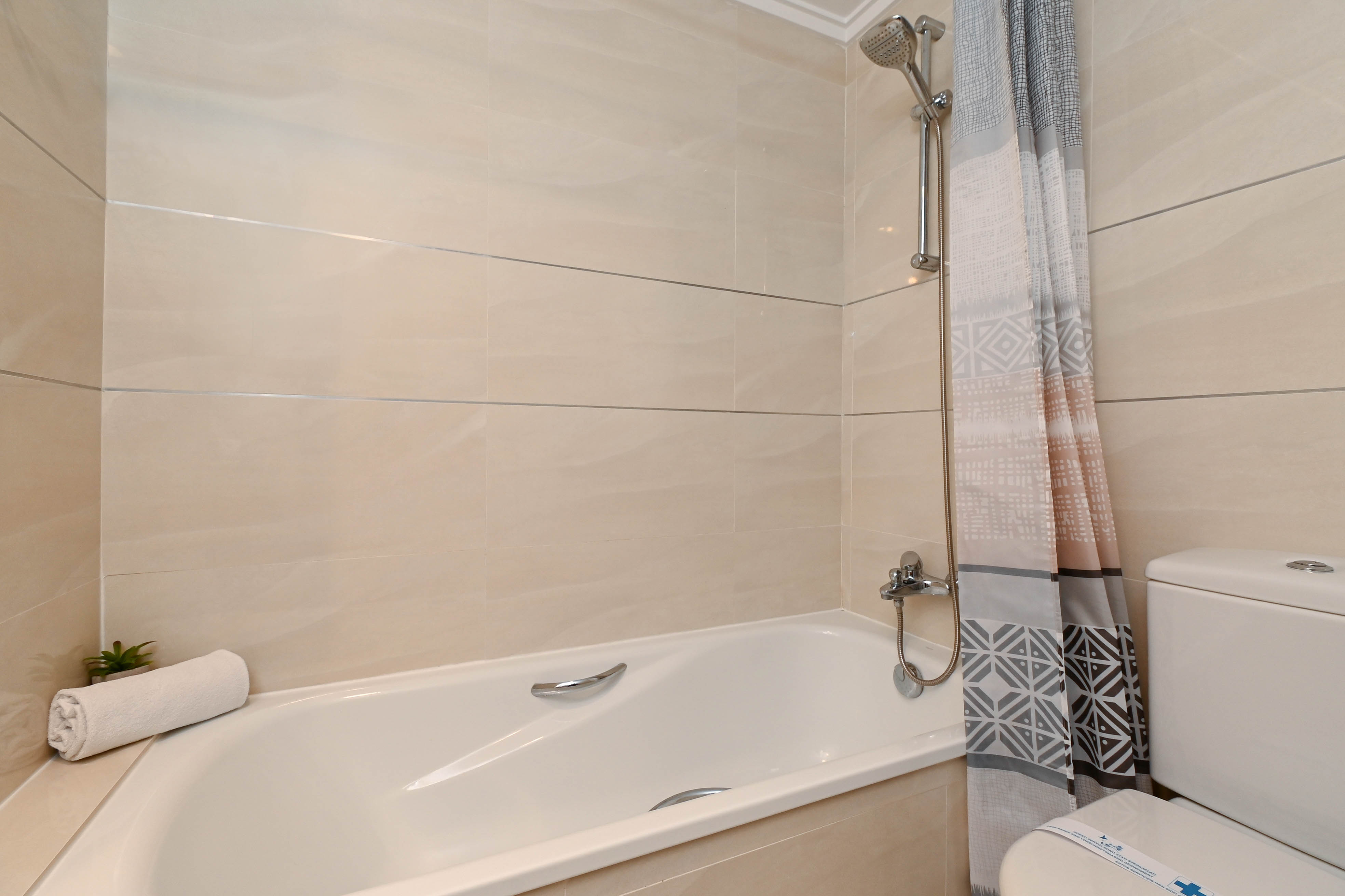 Cuarto de baño funcional con bañera, grifería cromada y paredes revestidas en azulejos beige con detalles horizontales. Incluye cortina con motivos geométricos.