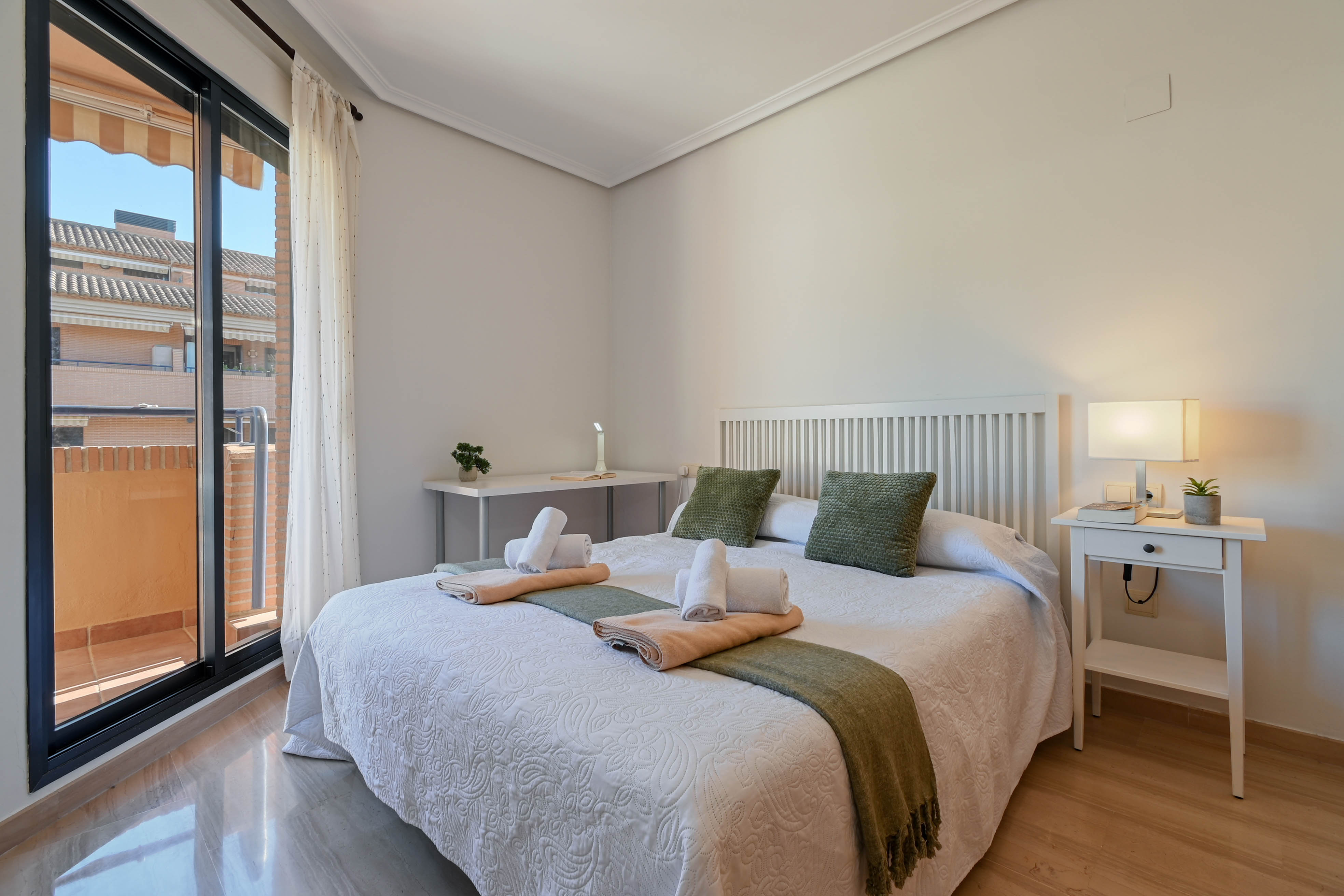Dormitorio luminoso con cama doble, cabecero de listones blancos, zona de escritorio y acceso directo a un balcón privado mediante puertas correderas.