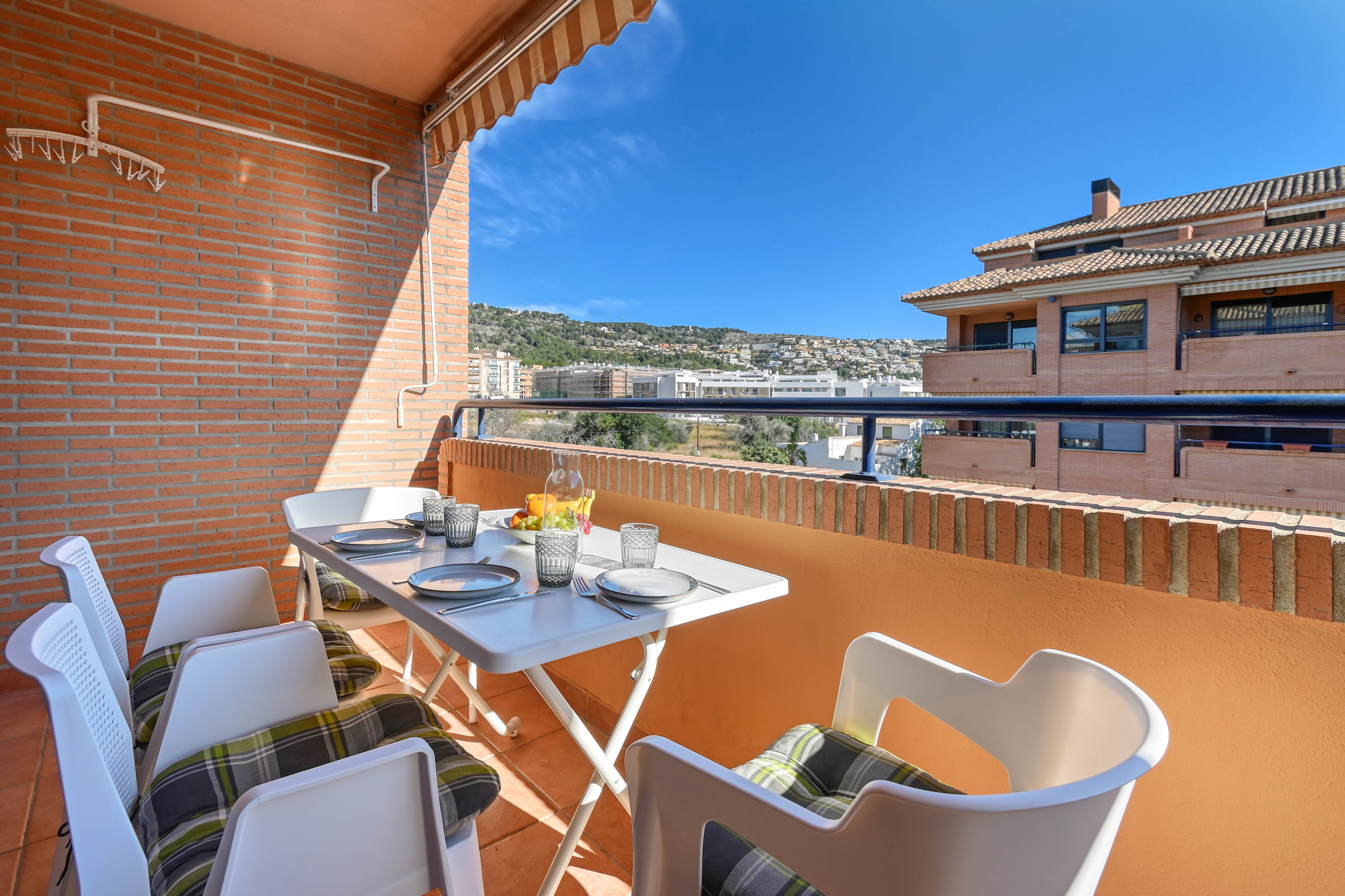 Terraza privada con mesa de comedor exterior, toldo extensible y paredes de ladrillo visto. Ofrece vistas despejadas hacia las colinas y el entorno residencial de Jávea.