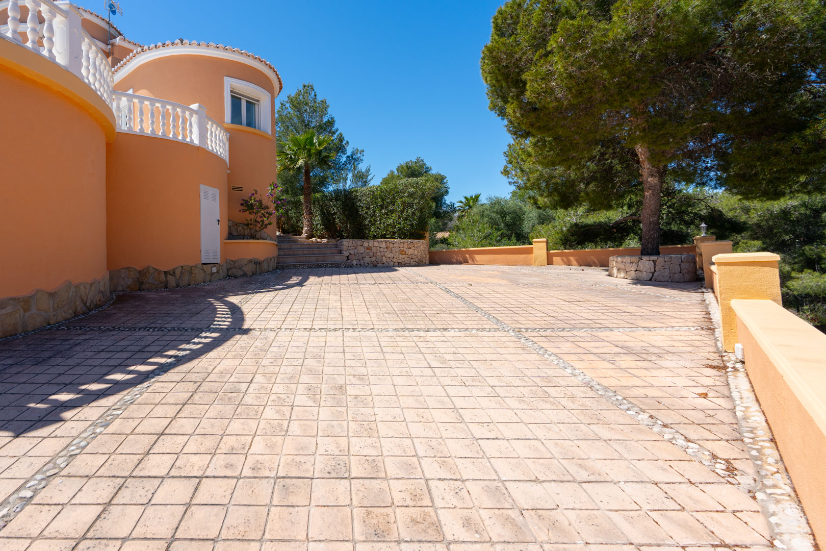 Amplia terraza exterior pavimentada con baldosas y detalles en piedra, rodeada de muros curvos en tono terracota y vegetación mediterránea autóctona.