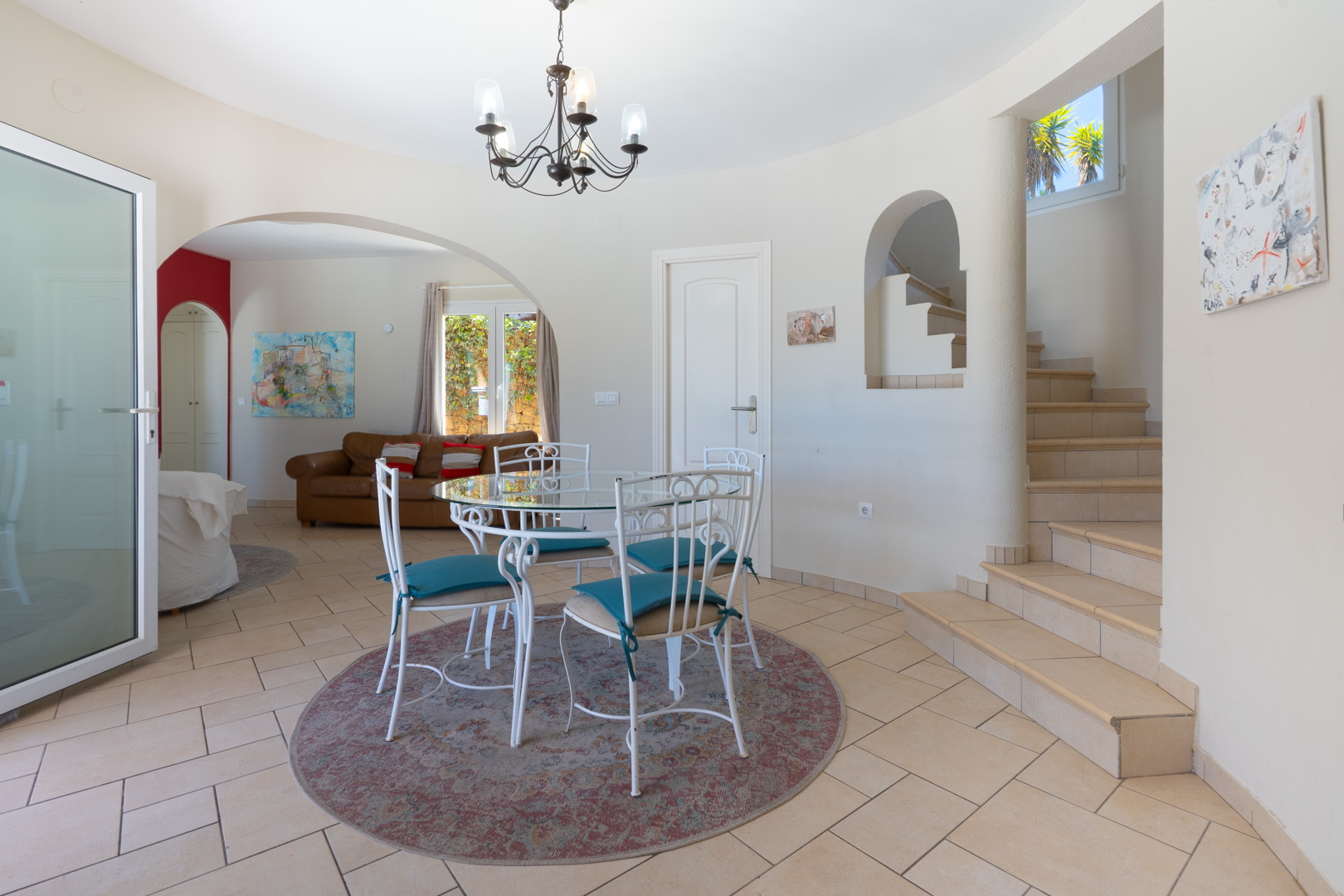 Comedor con mesa de cristal y sillas de hierro forjado blanco sobre alfombra circular. Incluye acceso a la planta superior por escalera de gres y vista al salón.