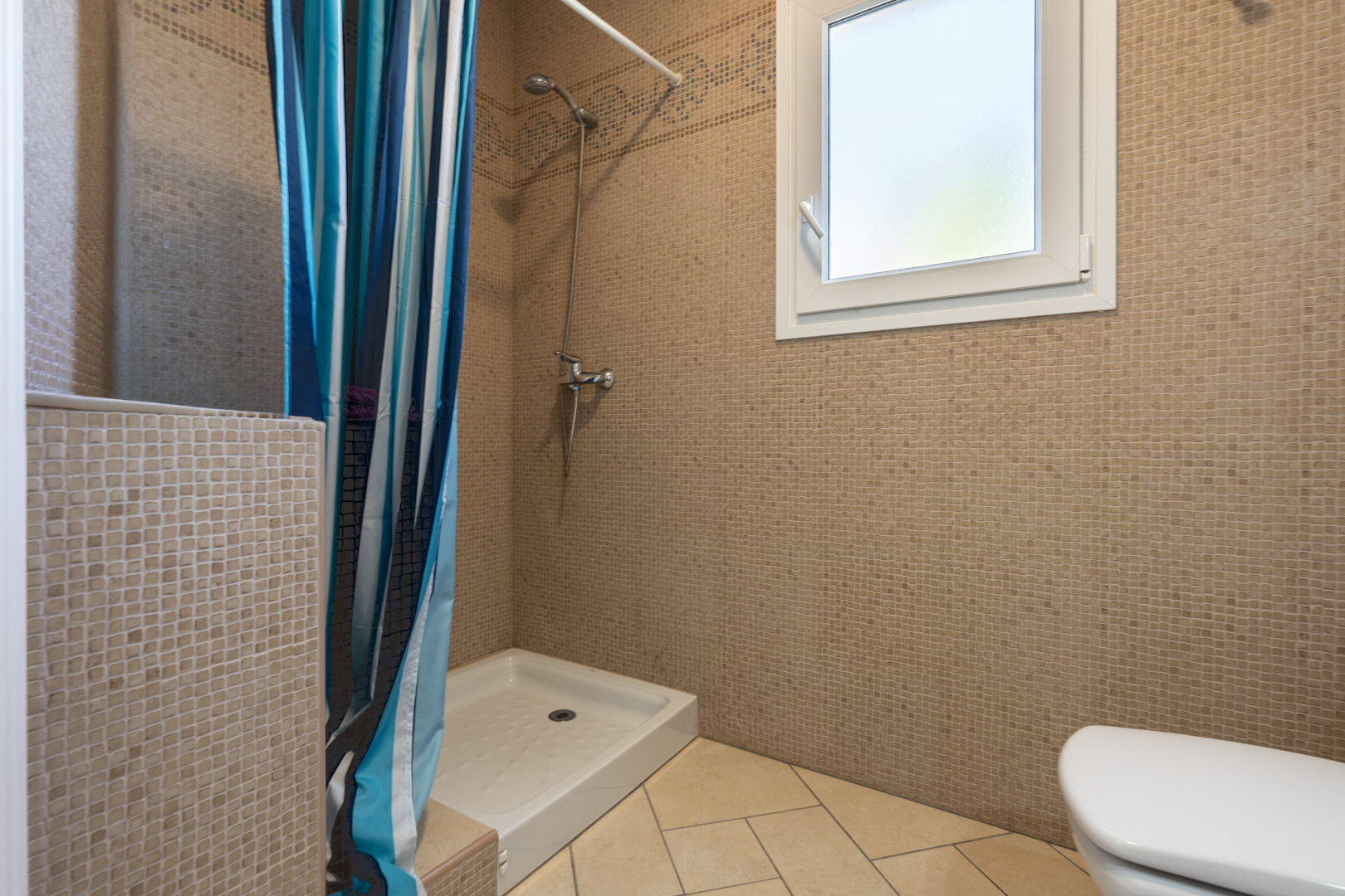 Cuarto de baño con paredes de mosaico marrón, plato de ducha blanco con cortina a rayas y ventana de PVC con cristal traslúcido para mayor privacidad.