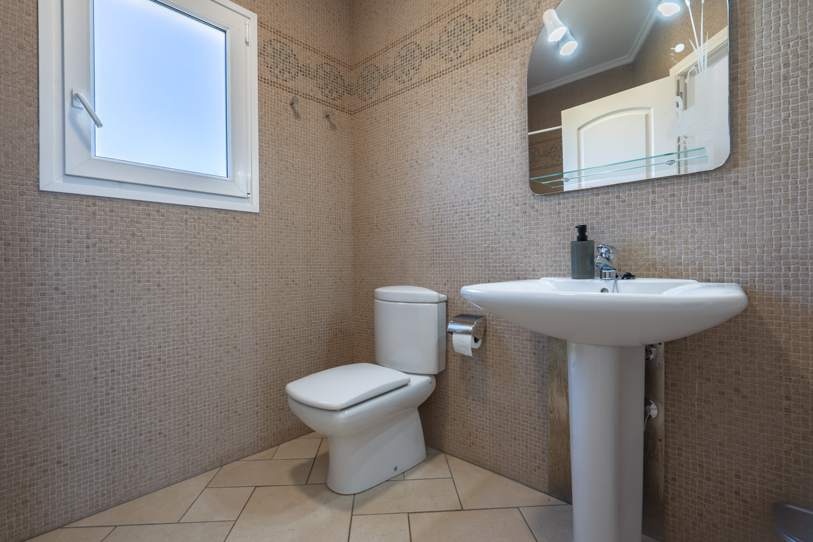 Cuarto de baño con paredes revestidas de mosaico beige, lavabo de pedestal, inodoro y ventana de PVC que aporta luz natural.
