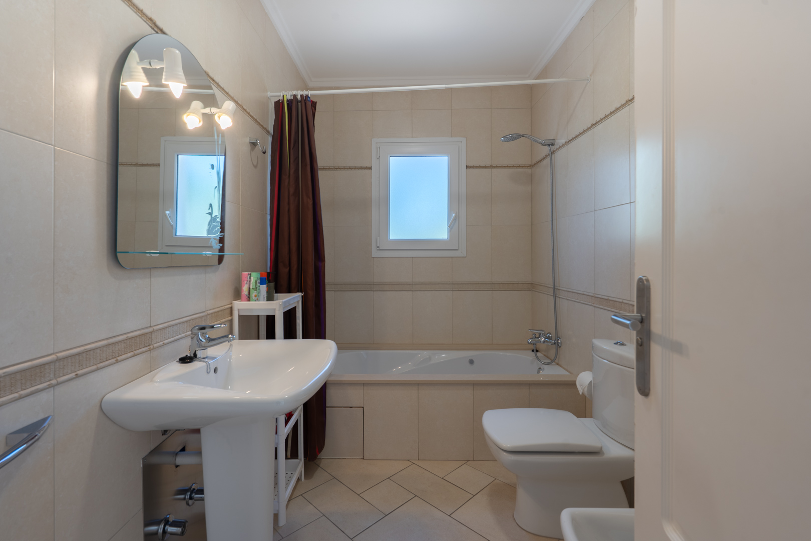 Cuarto de baño con revestimiento cerámico beige, bañera encastrada, lavabo de pedestal y espejo arqueado con iluminación integrada sobre ventana de PVC.