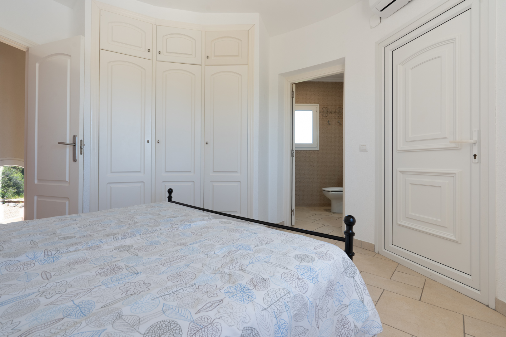 Dormitorio con armarios empotrados lacados en blanco, suelos cerámicos y acceso directo a un baño en suite con revestimiento de gresite.