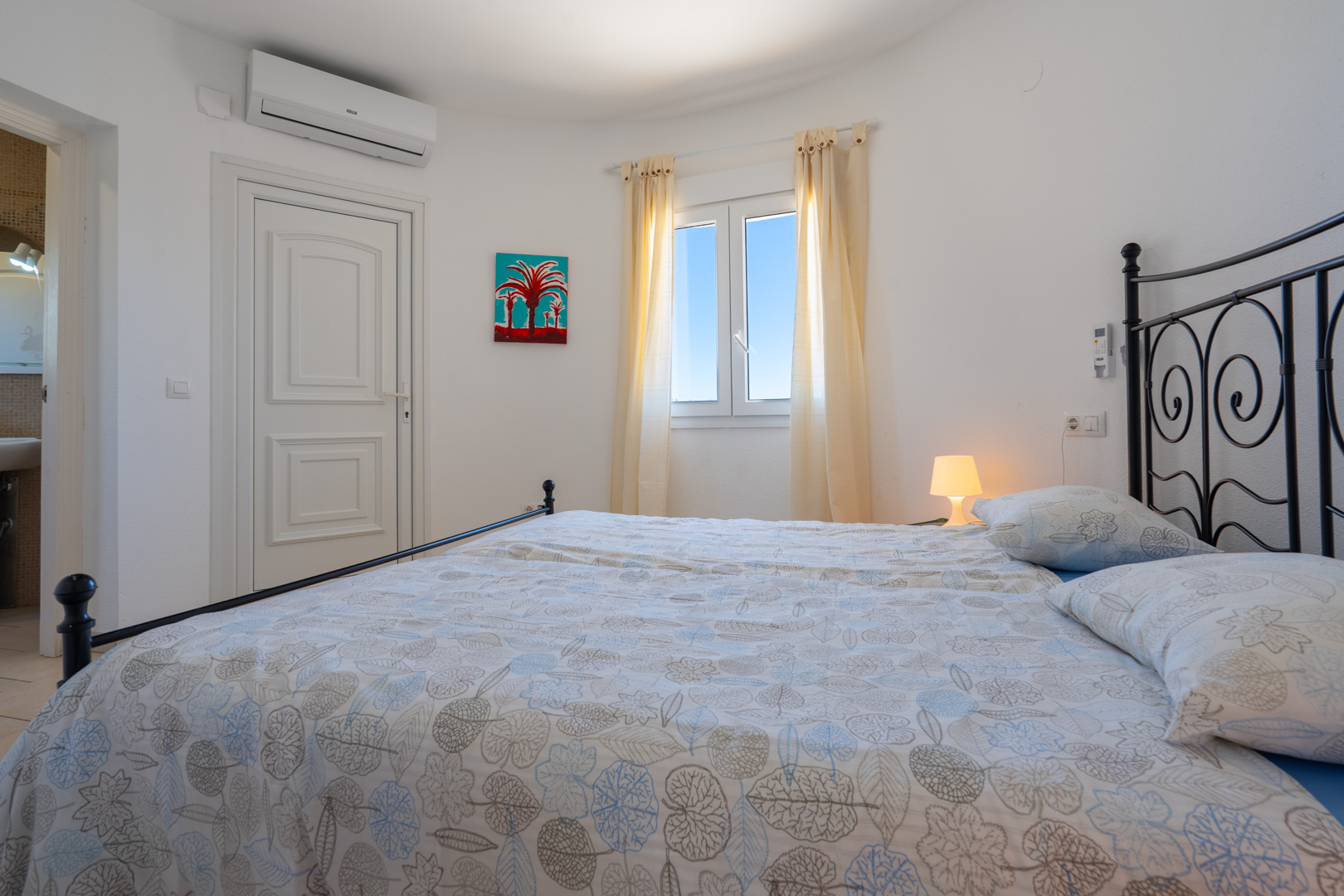 Dormitorio luminoso con cama de forja negra, aire acondicionado, ventana de PVC y acceso directo a un baño privado funcional.