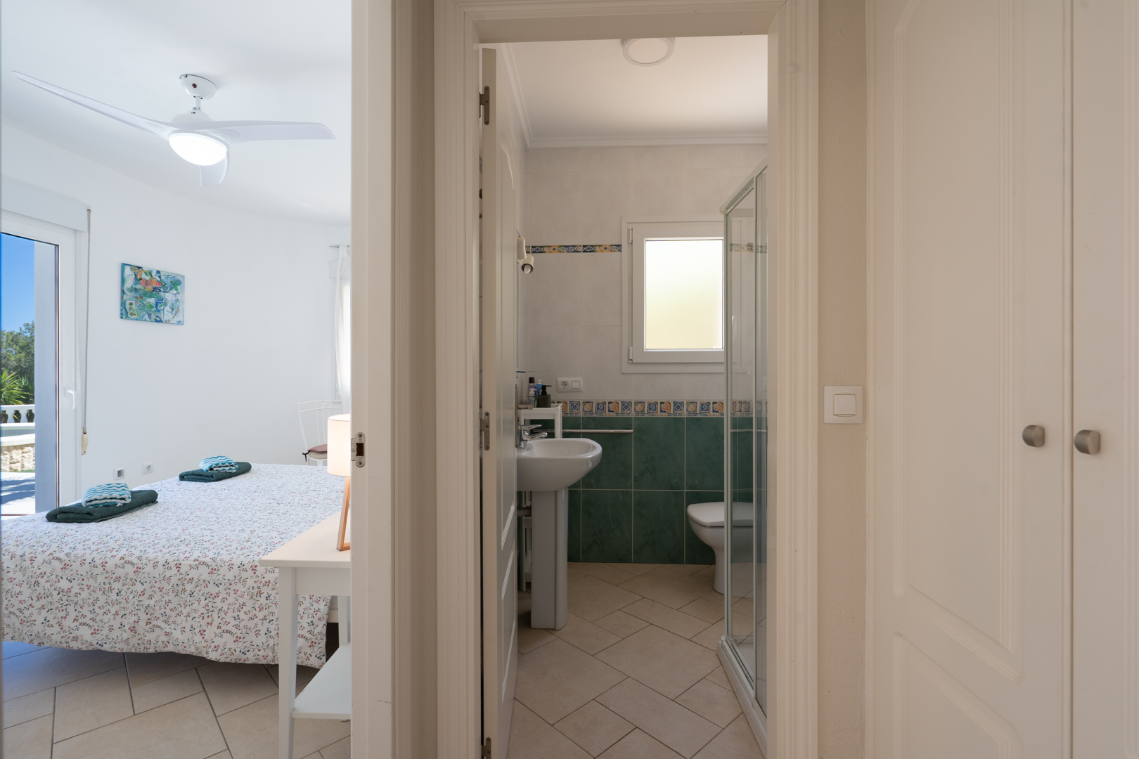 Dormitorio luminoso con ventilador de techo y acceso directo al exterior, ubicado junto a un baño con ducha y armarios empotrados en el pasillo.