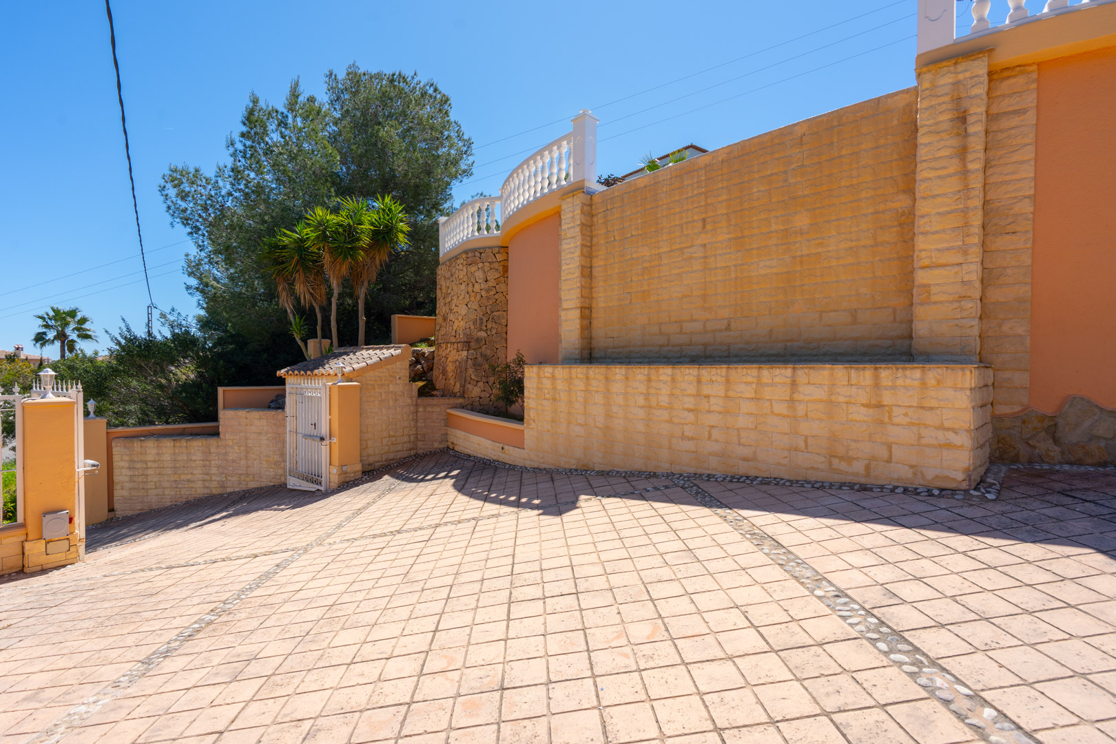 Entrada principal con amplio camino pavimentado en patrón de diamante, muros de piedra natural y terraza superior con balaustrada blanca bajo el cielo despejado de Jávea.