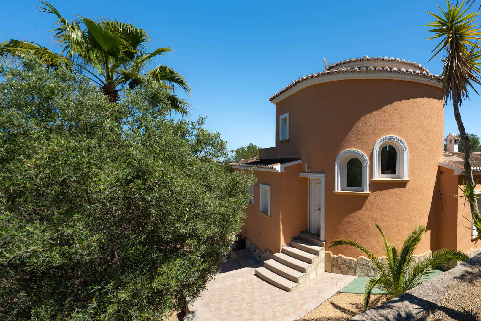 Exterior de la villa con fachada en tono tierra, destacando una estructura circular con ventanas de arco y acceso principal mediante escalones de piedra.