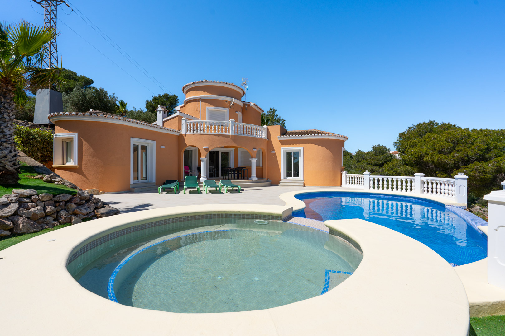 Exterior de la villa con piscina de diseño curvo, amplia terraza pavimentada y fachada en tonos tierra con detalles arquitectónicos redondeados y balaustradas blancas.