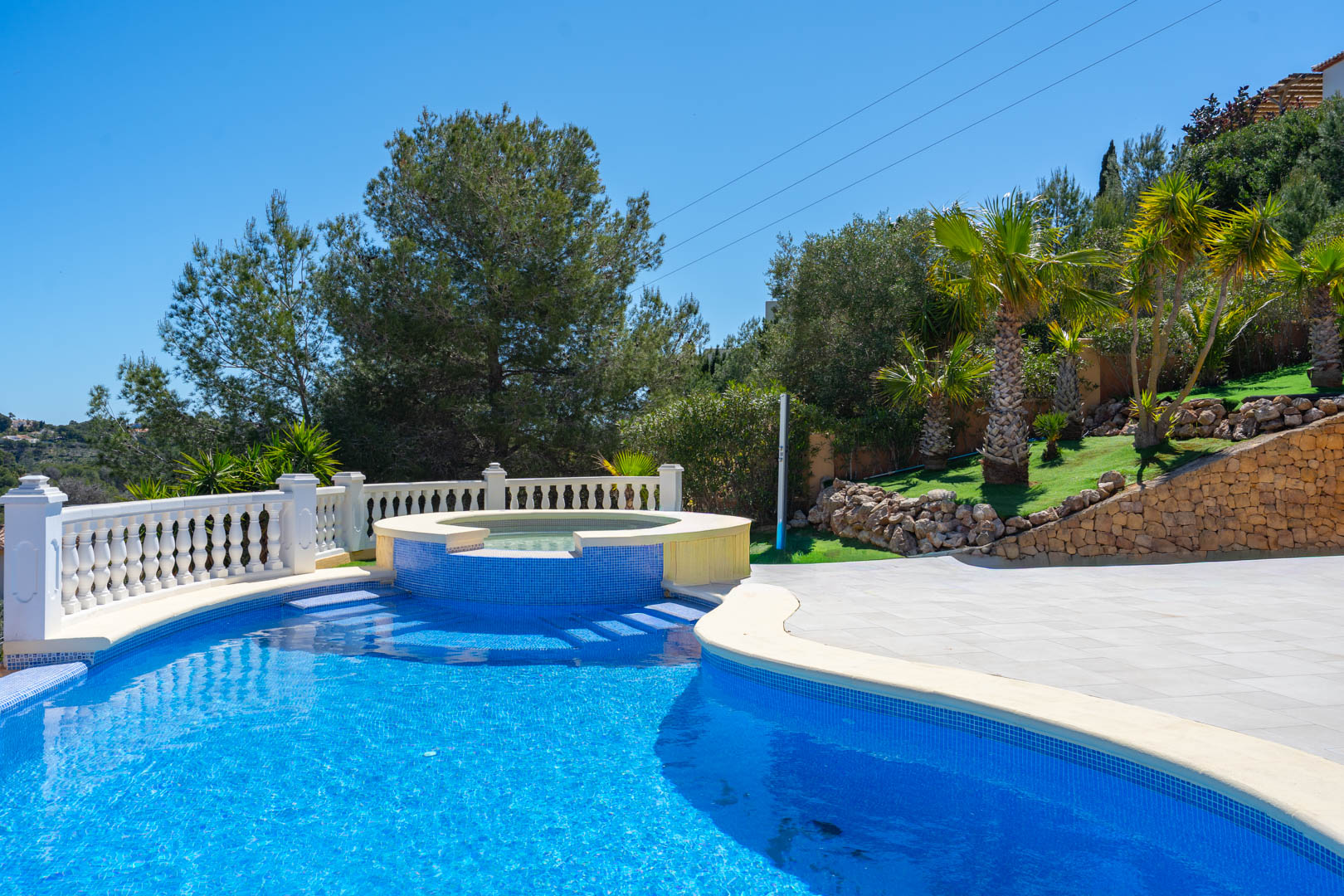 Piscina de 10x5 m con jacuzzi integrado y azulejos azules, rodeada por una terraza pavimentada, balaustrada blanca y jardín mediterráneo con palmeras.