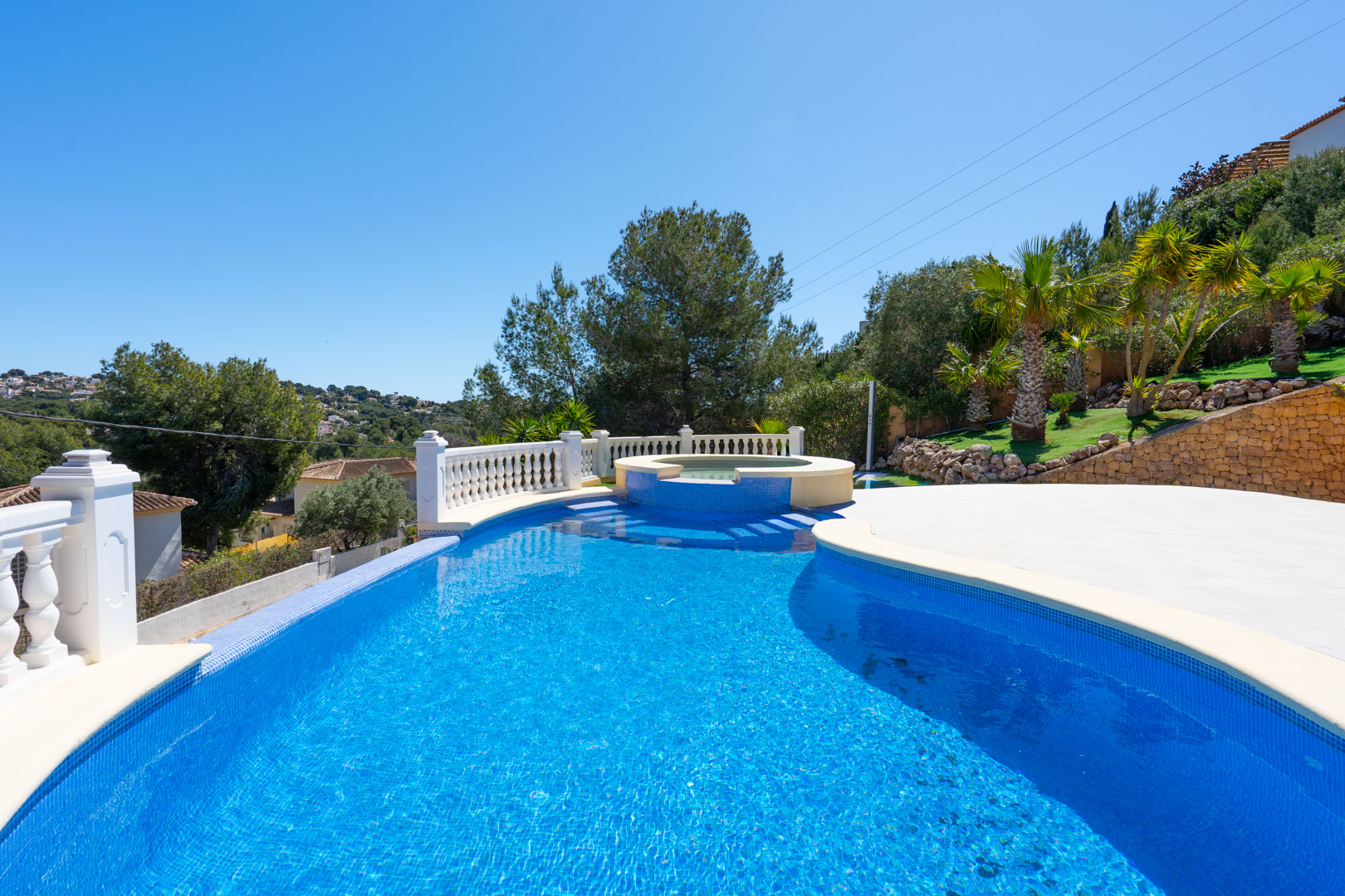 Piscina de gresite azul con jacuzzi integrado, rodeada por una terraza pavimentada, balaustrada blanca y jardín con palmeras y muros de piedra natural.