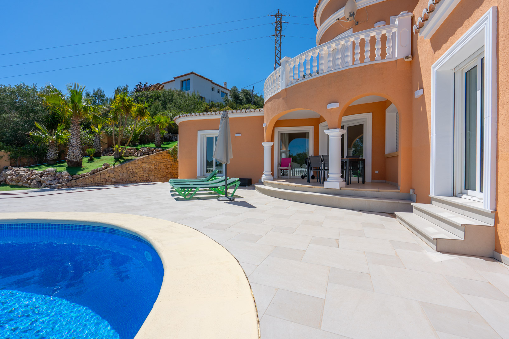 Terraza exterior con piscina curva y porche de arcos blancos. La fachada de tonos cálidos se complementa con balaustradas clásicas y un jardín mediterráneo con palmeras.