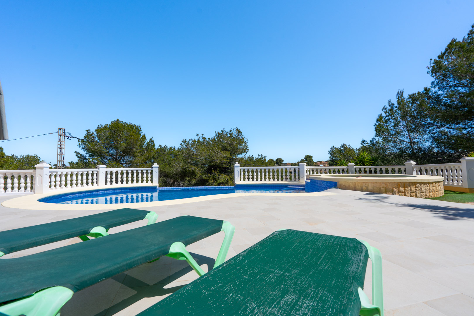 Terraza exterior con piscina de azulejos azules, balaustrada blanca y zona de jacuzzi integrada, rodeada de pinos y tumbonas sobre suelo de piedra clara.
