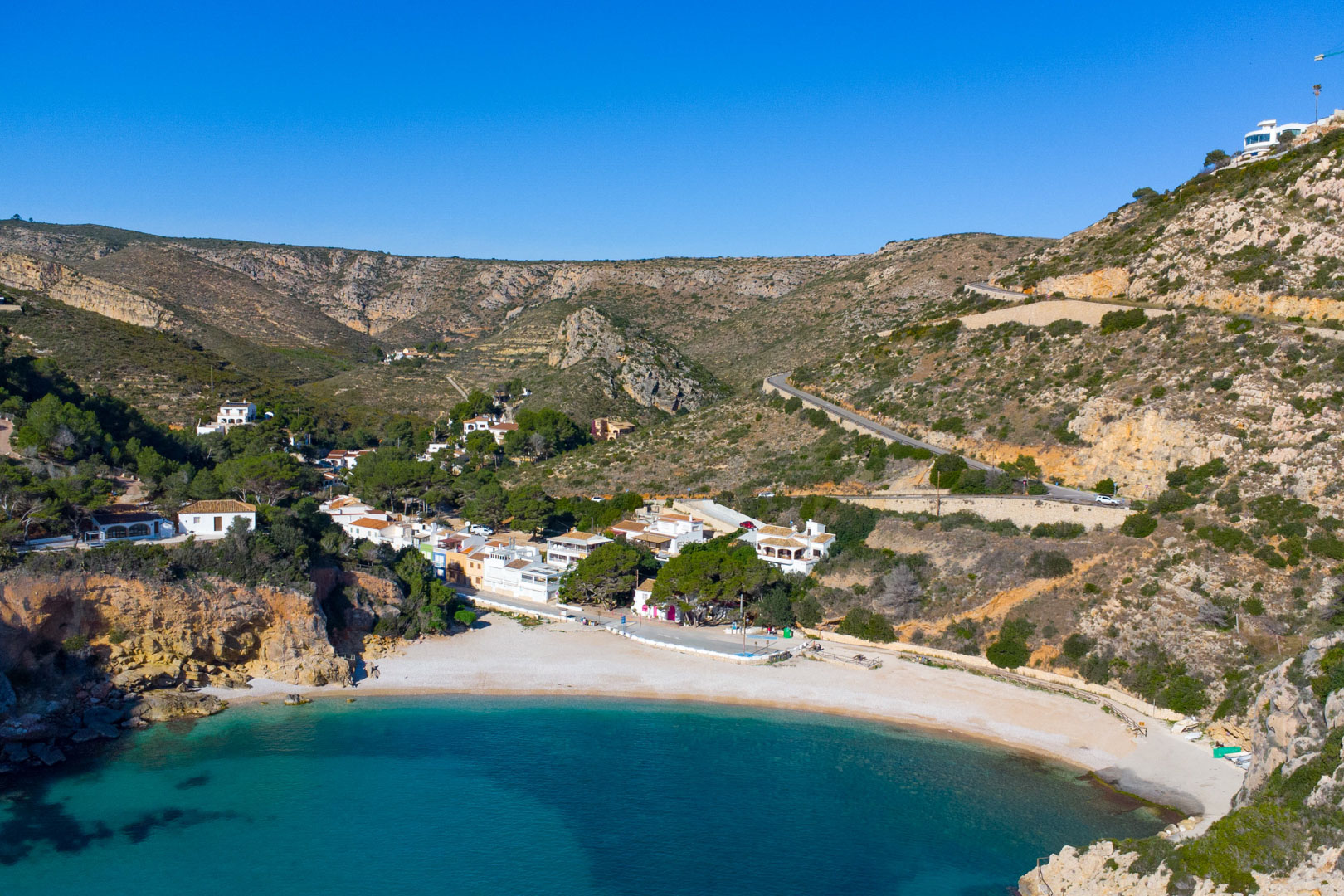 Vista aérea de la Cala Granadella en Jávea, mostrando sus aguas cristalinas, la playa de guijarros y el entorno montañoso con acceso por carretera sinuosa.
