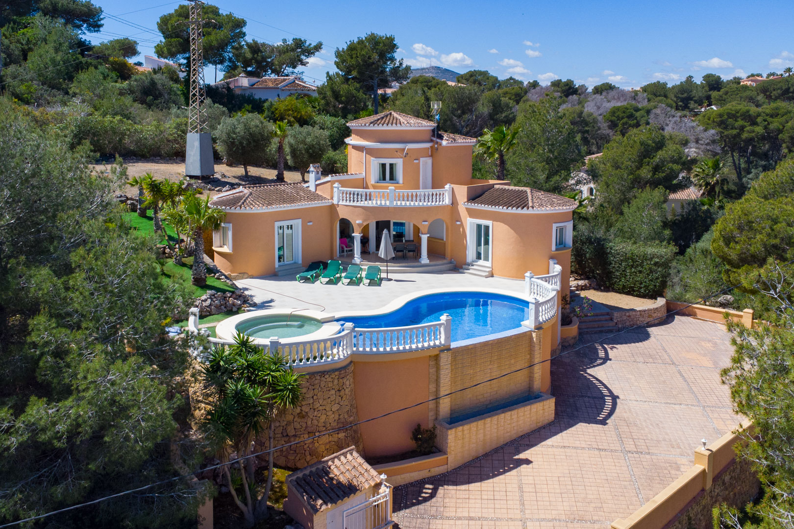 Vista aérea de la villa mediterránea con piscina curva, jacuzzi integrado y balaustradas blancas, rodeada de pinos y una amplia zona pavimentada.