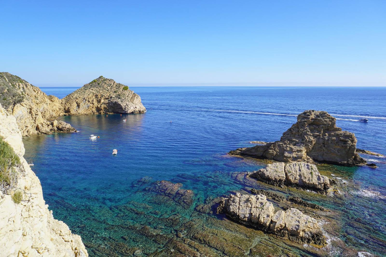 Vista panorámica de la costa de Jávea con aguas cristalinas de color turquesa, formaciones rocosas naturales y pequeñas embarcaciones fondeadas bajo un cielo despejado.