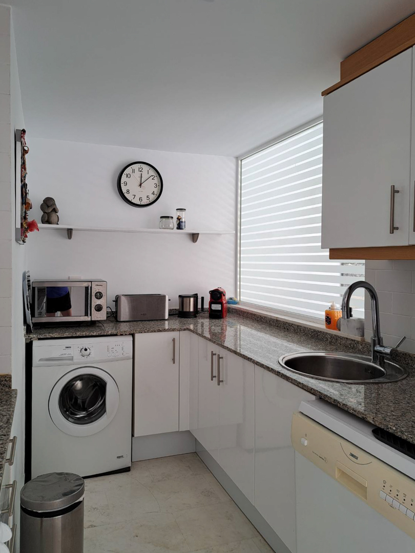 Cocina funcional en forma de L con encimera de granito, armarios blancos, lavadora integrada y una amplia ventana que proporciona abundante luz natural.