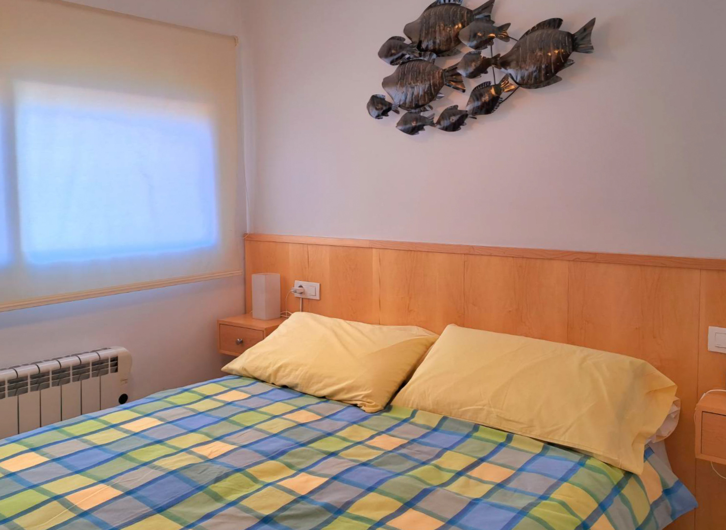 Dormitorio con cabecero de madera integrado, mesitas de noche a juego y decoración de pared metálica. Incluye radiador eléctrico y ventana con estor.