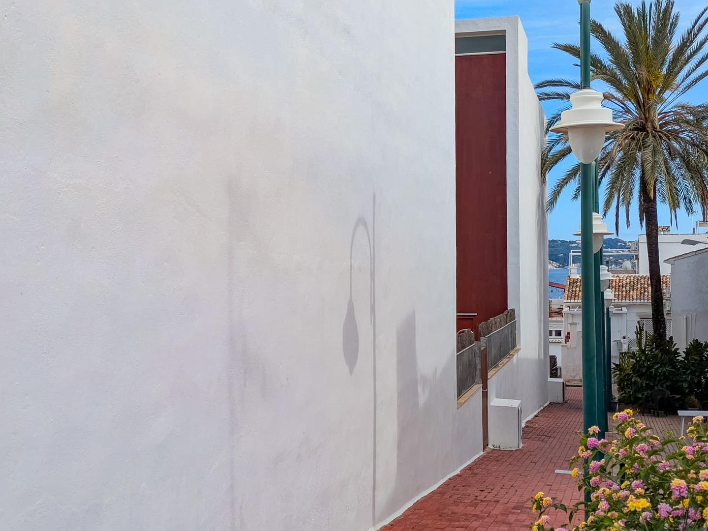 Exterior del edificio con fachada blanca y camino peatonal de ladrillo que ofrece vistas parciales al mar y vegetación mediterránea.