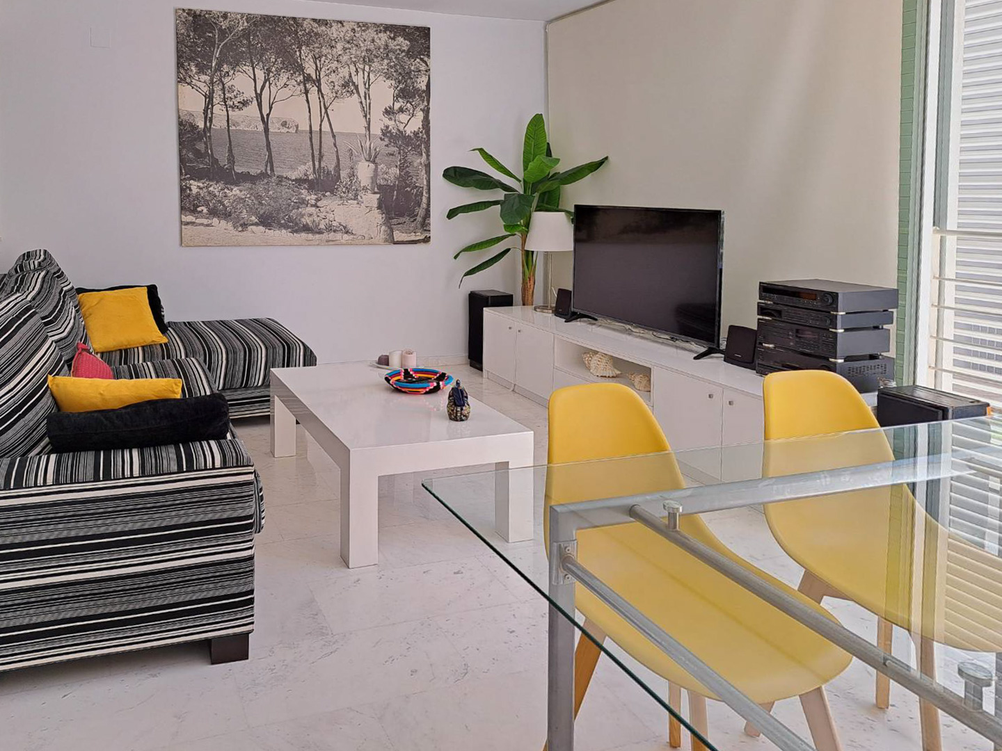 Salón-comedor con suelos de mármol, sofá de rayas y mesa de cristal con sillas amarillas. La estancia cuenta con un mueble de TV blanco y luz natural lateral.
